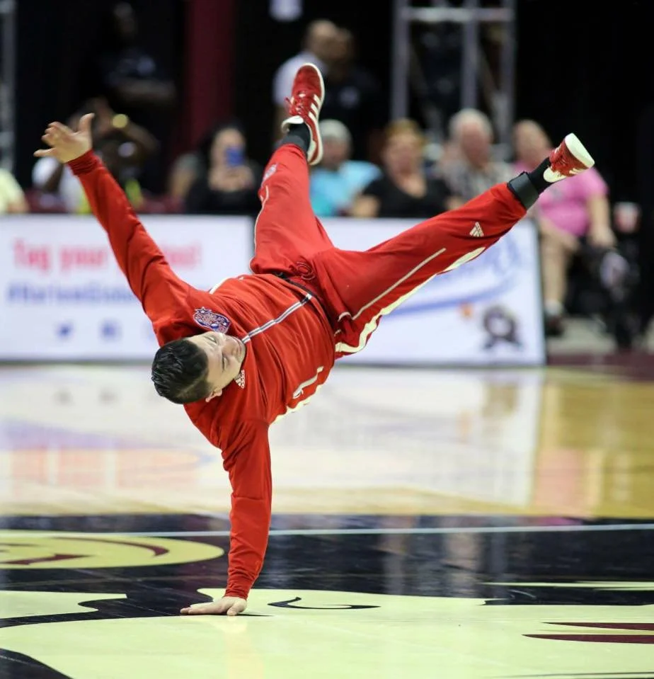 Timstuh performing a breakdance move on a basketball court, wearing a red tracksuit and red sneakers, with an audience in the background known for his World Tour and Travels.