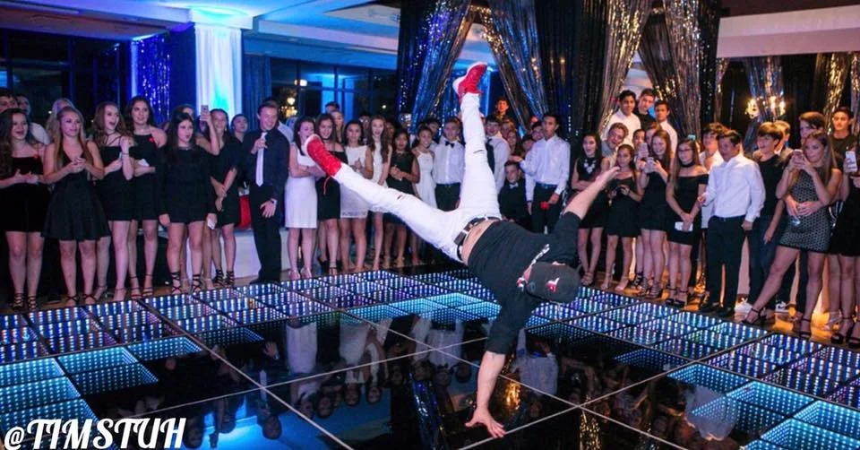 Timstuh in black and white pants and red shoes is doing a handstand on a glass dance floor at a party, with a crowd of young people watching and taking pictures in a decorated, festive room.
