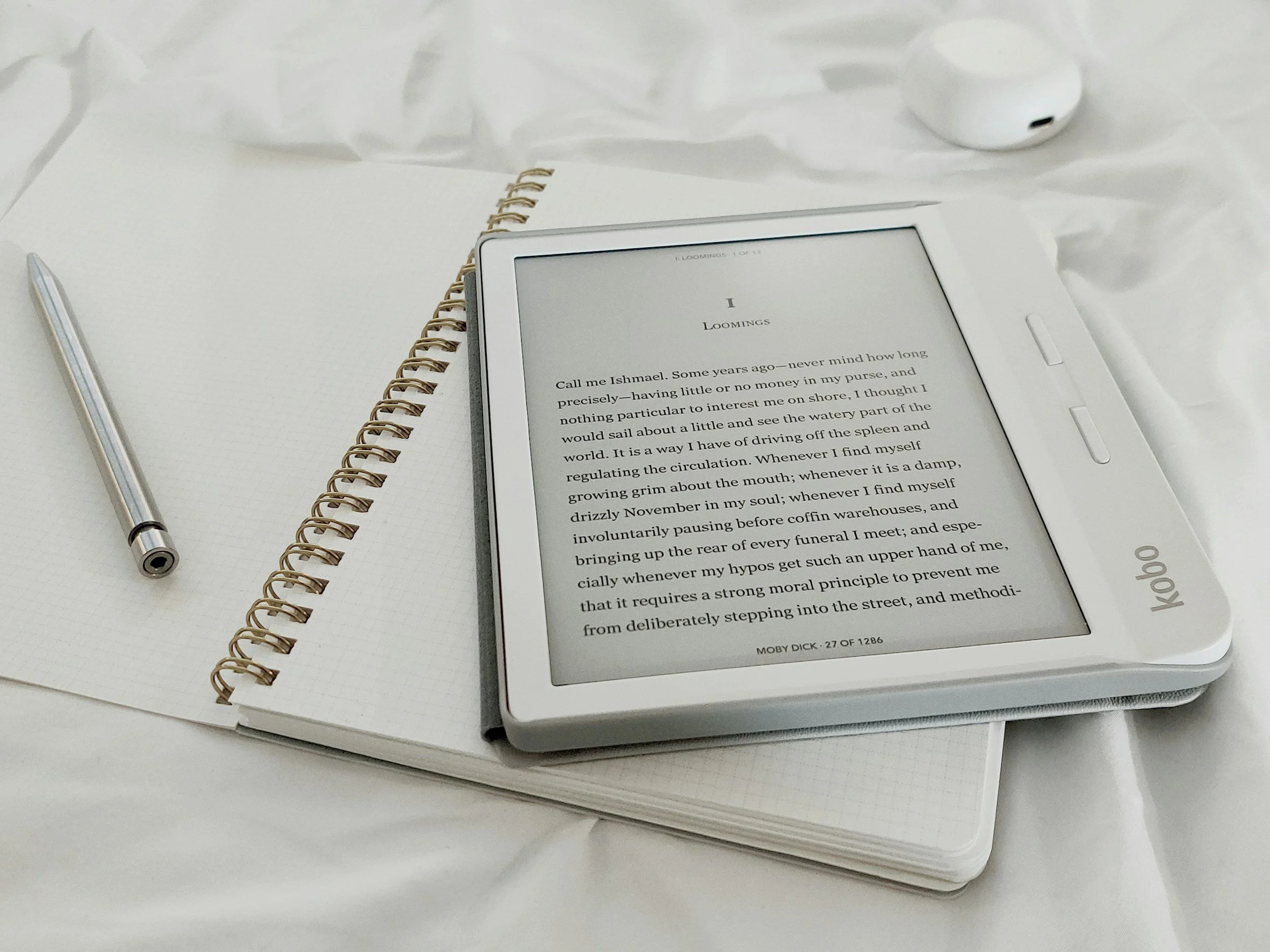An e-reader displaying text, placed on open notebooks with a pen nearby, and a wireless earbud in the background.