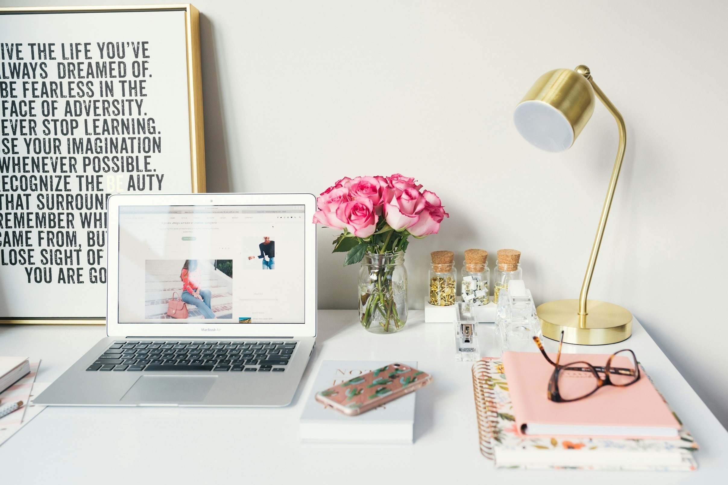 A white desk with a MacBook Air, a vase of pink roses, a pink phone with a cactus case, homemade notebooks, reading glasses, and a gold desk lamp. The workspace is decorated with framed art and small jars with gold and silver decorations.