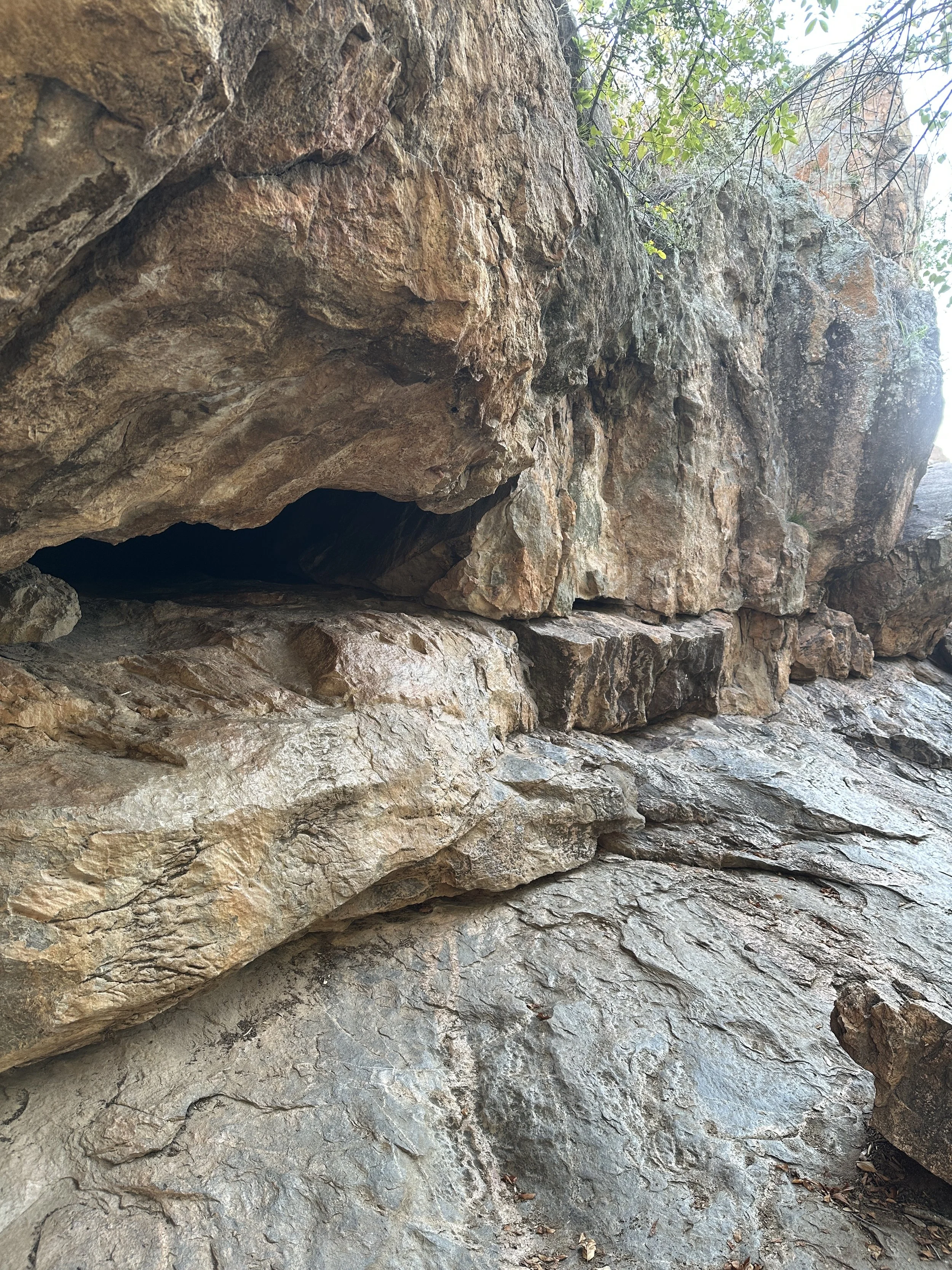 Large rocky formation with a dark cave opening, surrounded by layered rocks and some greenery at the top.