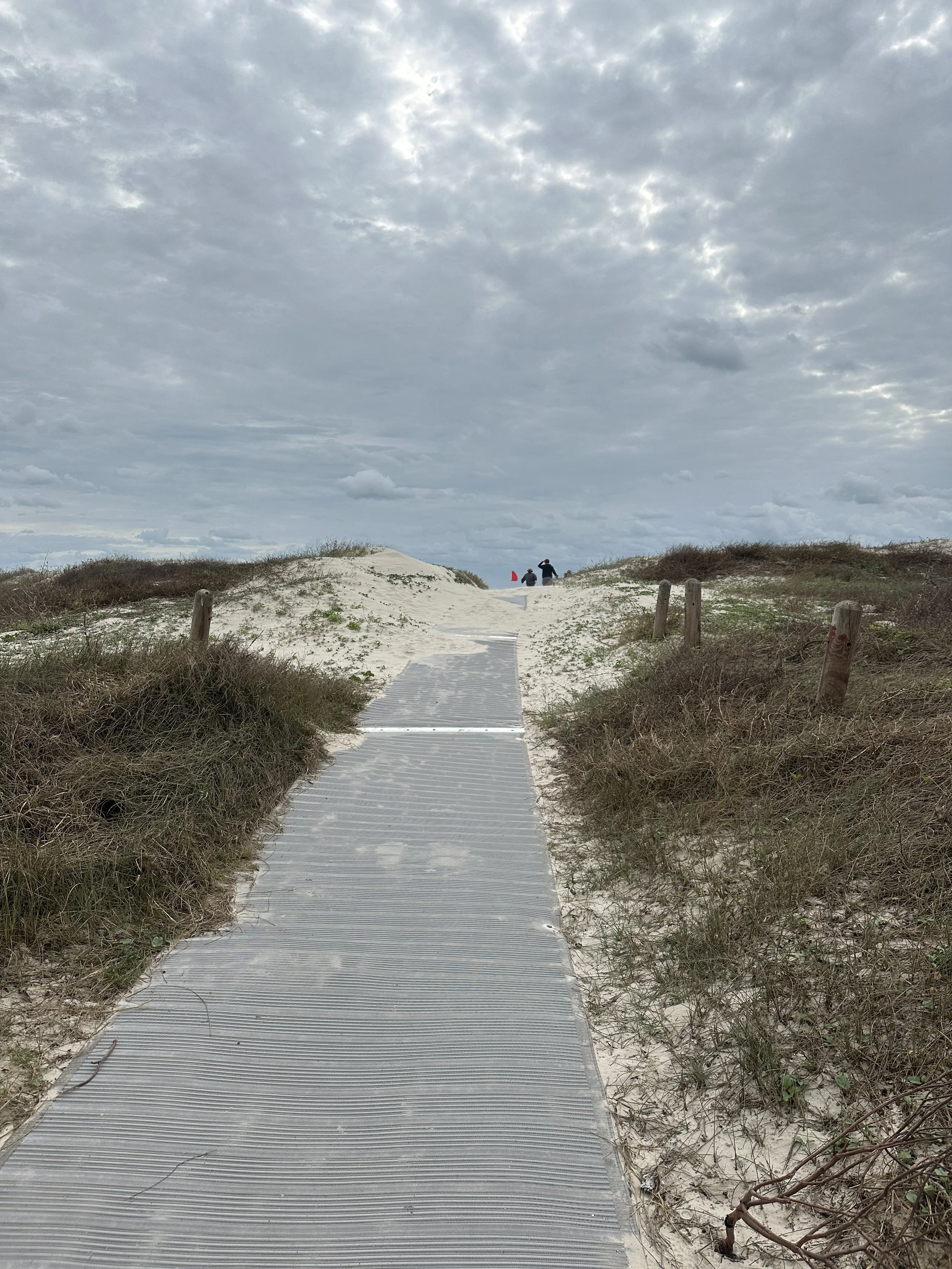 A sandy beach with a wooden pathway leading to the dunes, under a cloudy sky. Two people are walking near the top of the dunes in the distance.