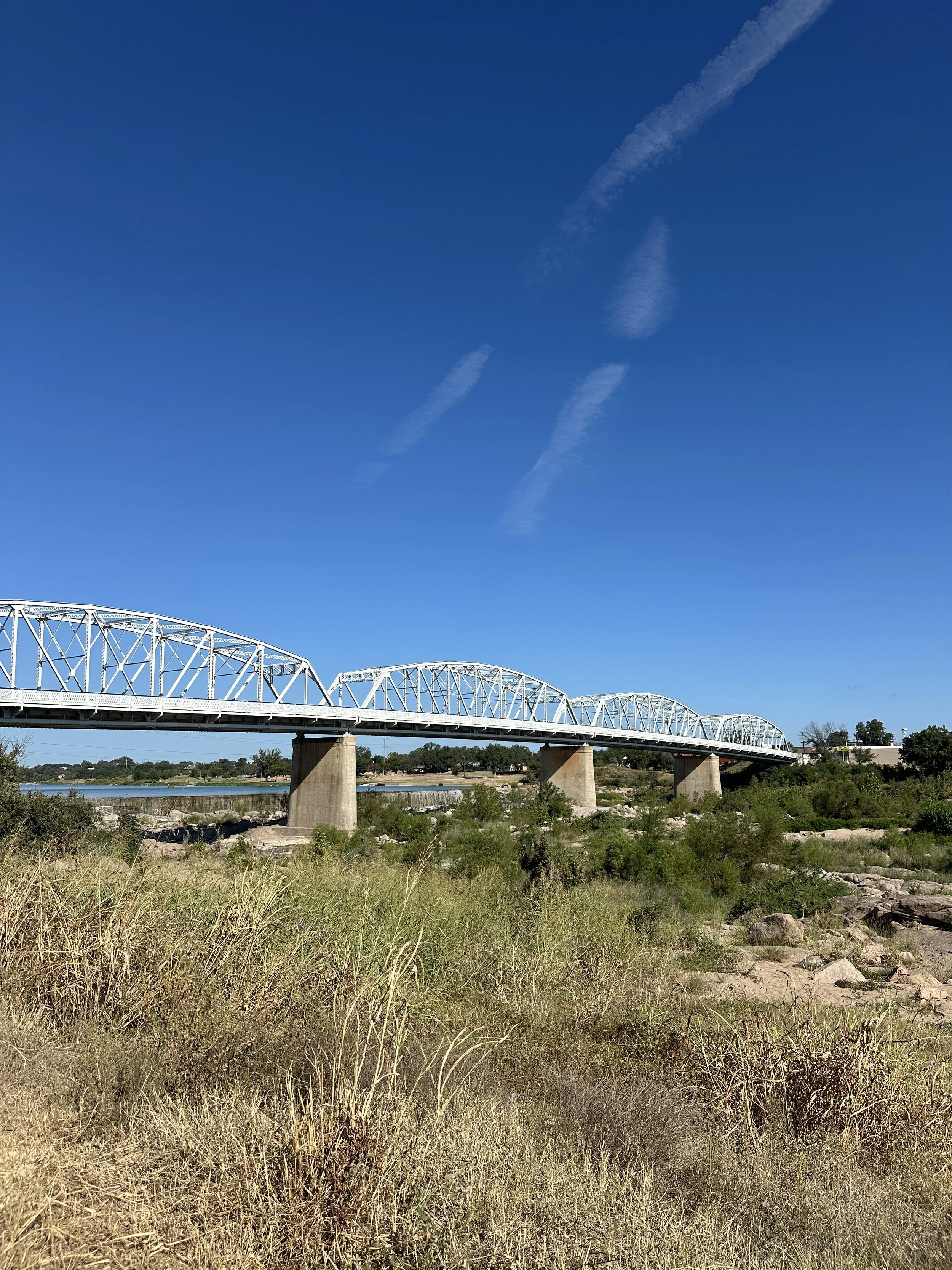 A white metal truss bridge spanning over a river, with a clear blue sky and wispy clouds in the background. There is dry grass and rocks in the foreground.