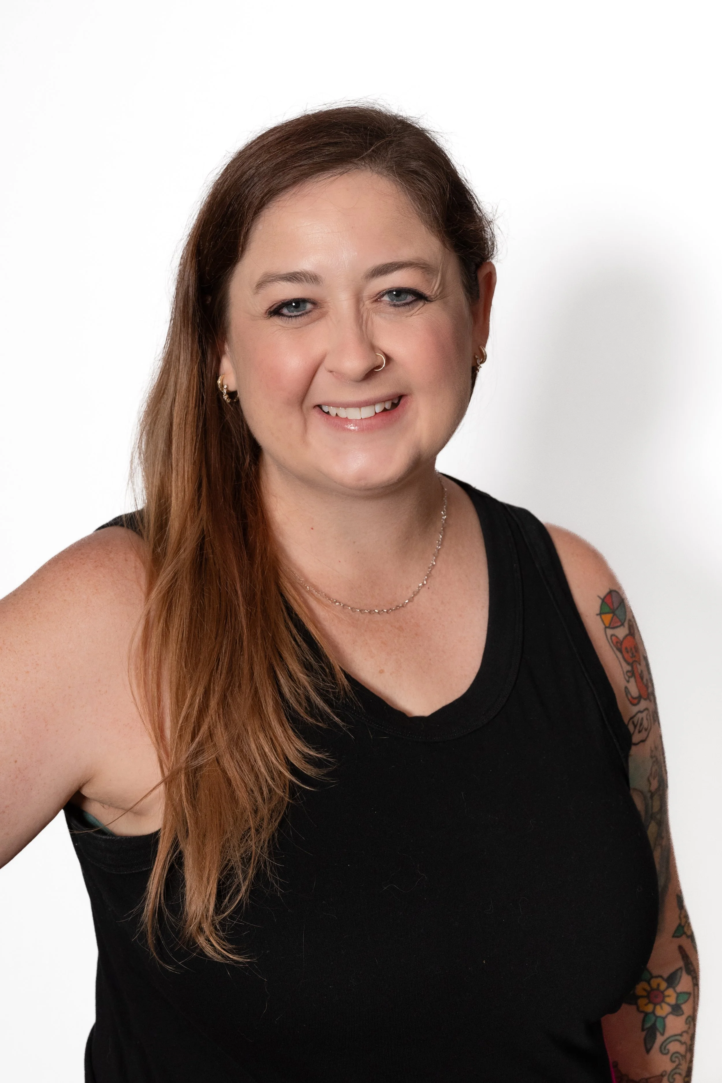 A smiling woman with long reddish hair, wearing a black sleeveless top, small hoop earrings, and a delicate necklace, standing against a white background.