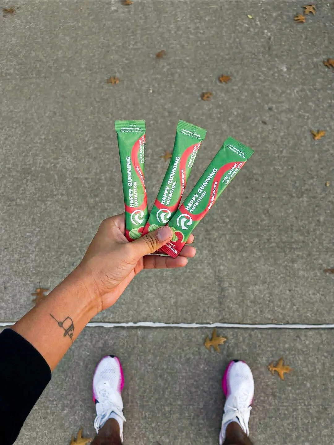 A person holding three green and red energy gel packs labeled 'Happy Running' on a concrete sidewalk with a few fallen leaves, wearing white athletic shoes with pink accents.