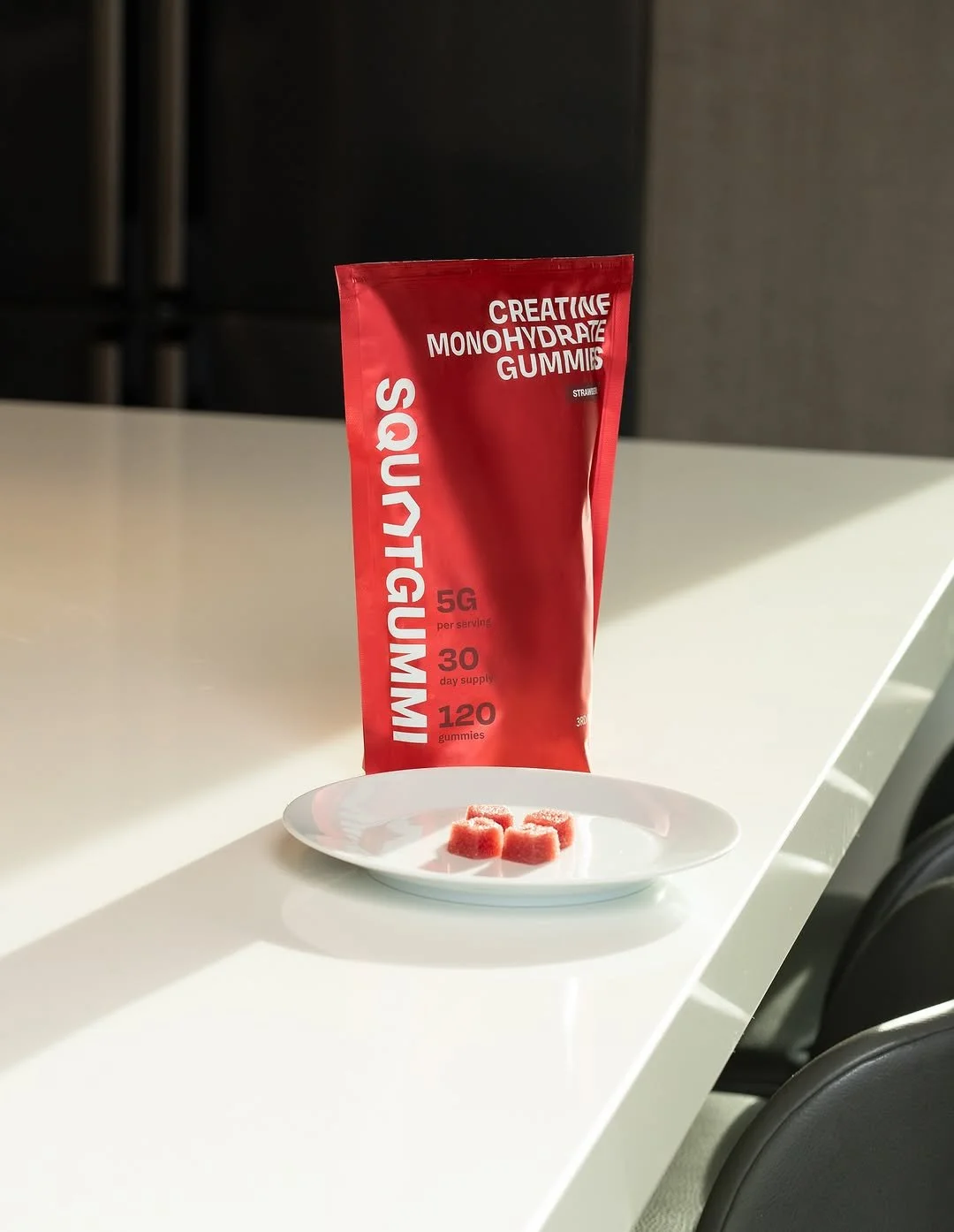 A red package labeled 'Creative Monohydrate Gummies' with nutritional info, placed on a white surface near a white plate with three pink gummy candies, in a room with dark furniture.