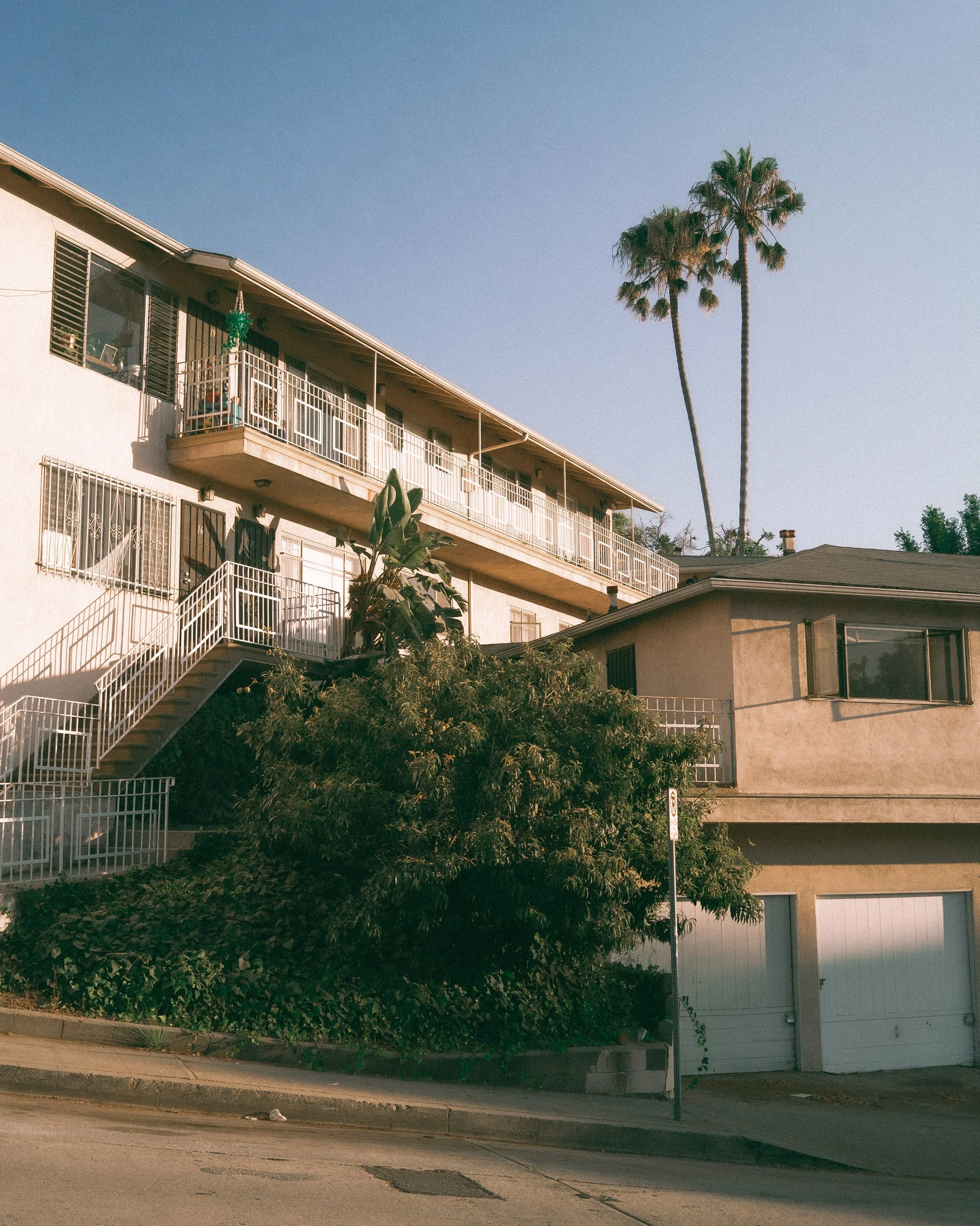 Apartment building with balconies, staircase, green foliage, palm trees, and a clear blue sky.