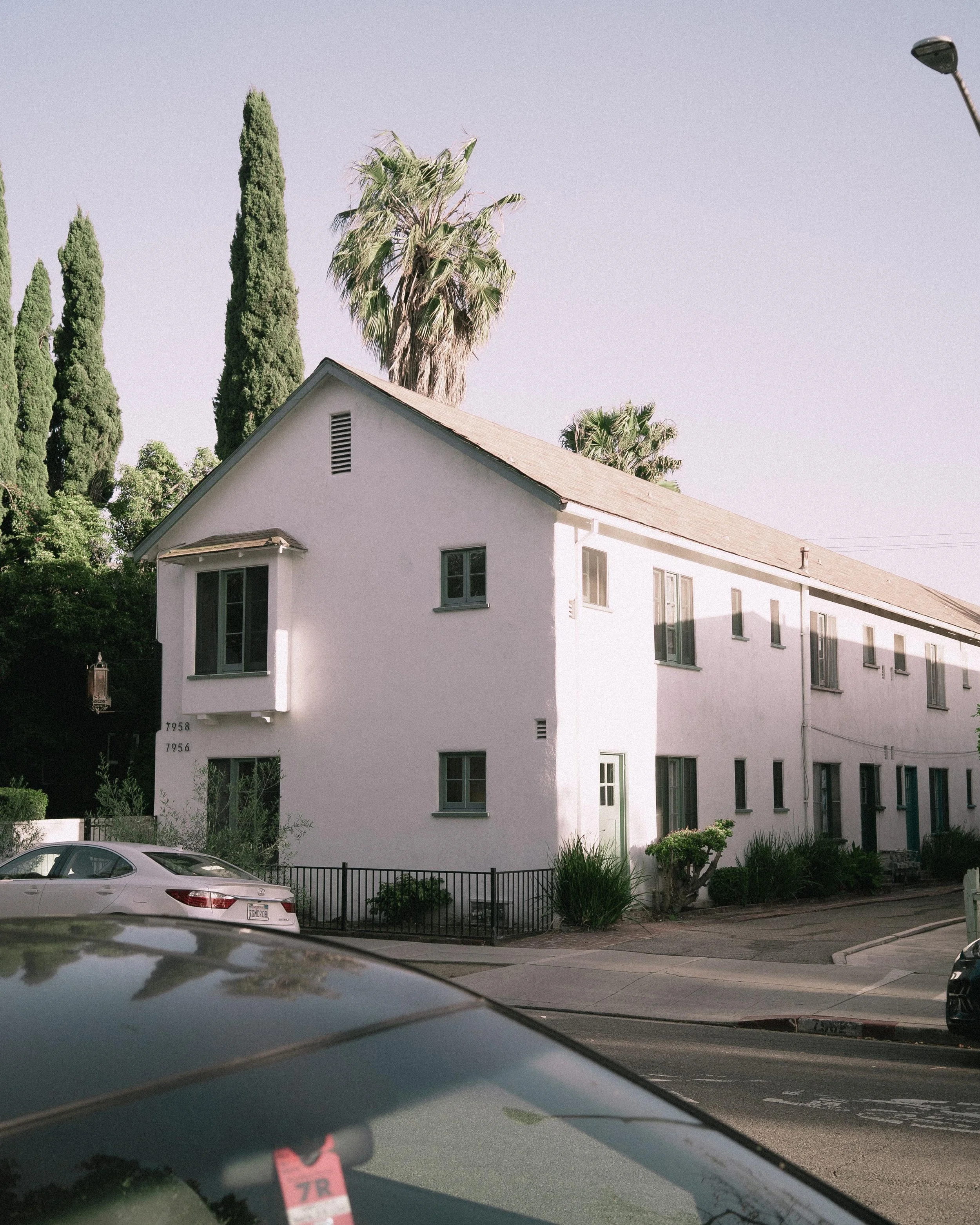 A white multi-story residential building with small windows and a green door, surrounded by plants and trees, including tall cypress and palm trees, with a silver car parked in front.
