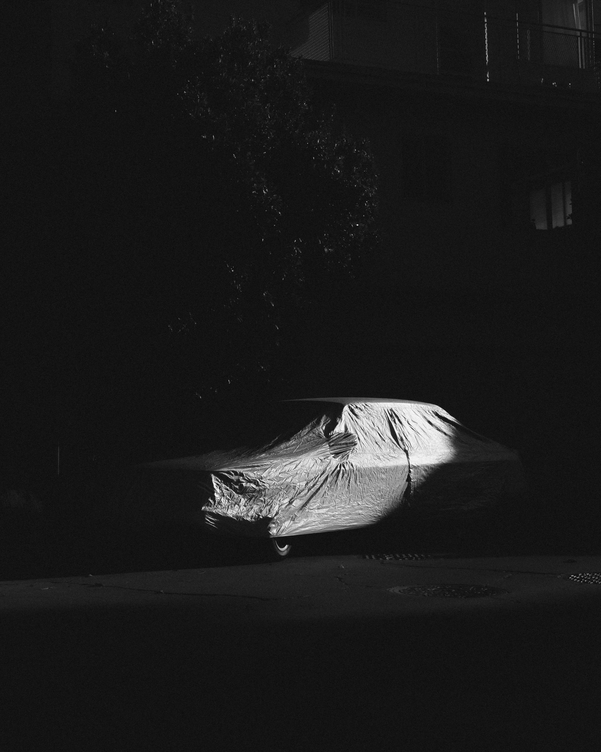 Car covered with a tarp parked on a street at night, with a tree and a building in the background.