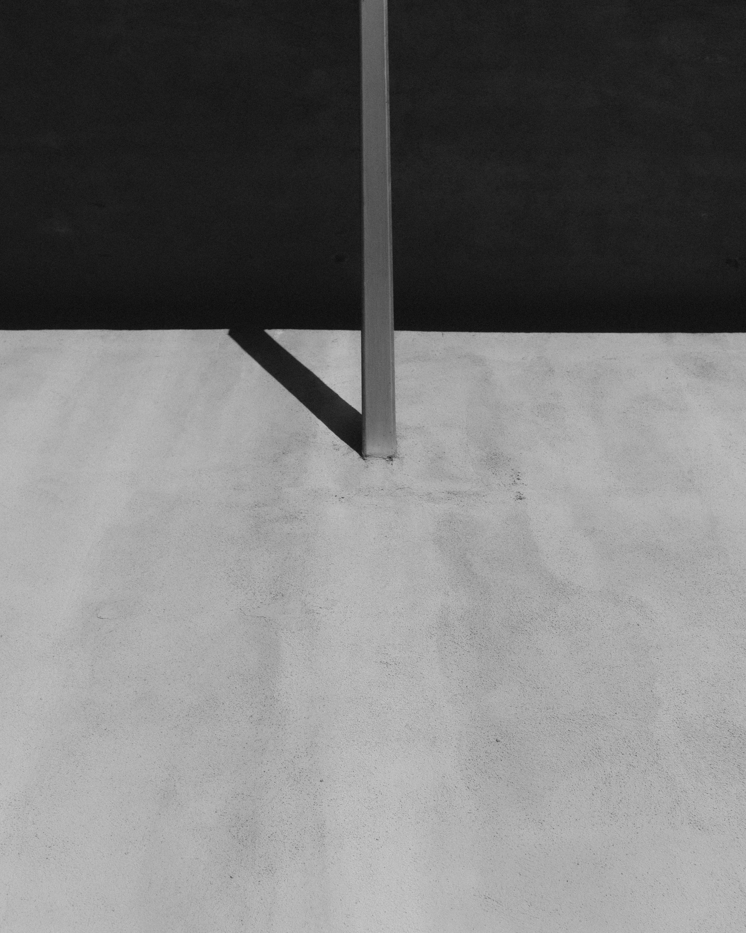 Black and white photo of a vertical metal pole casting a shadow on a flat surface, with a dark background.