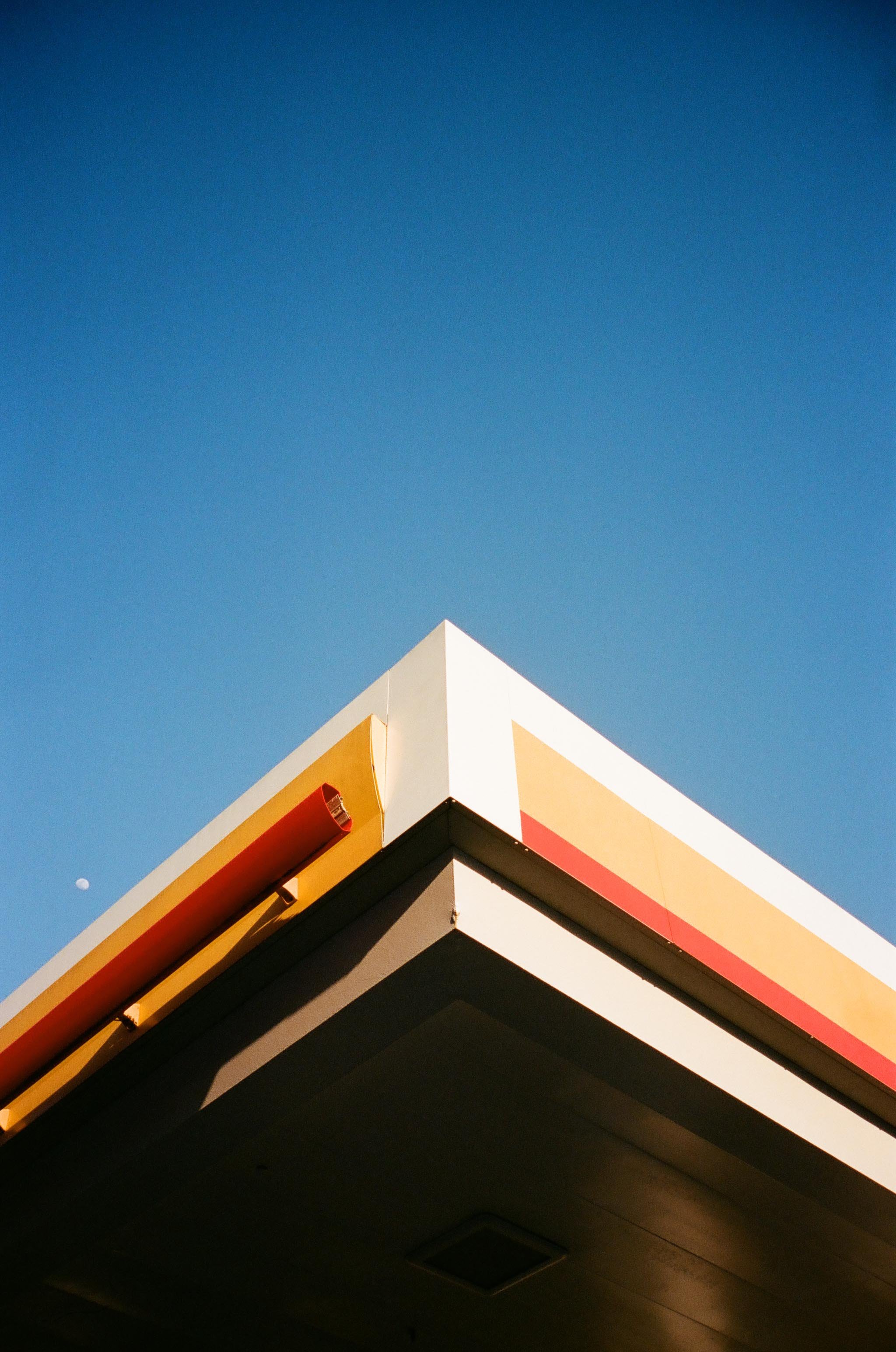 Close-up of the corner of a building with a colorful sign against a clear blue sky, with a small moon visible in the background.