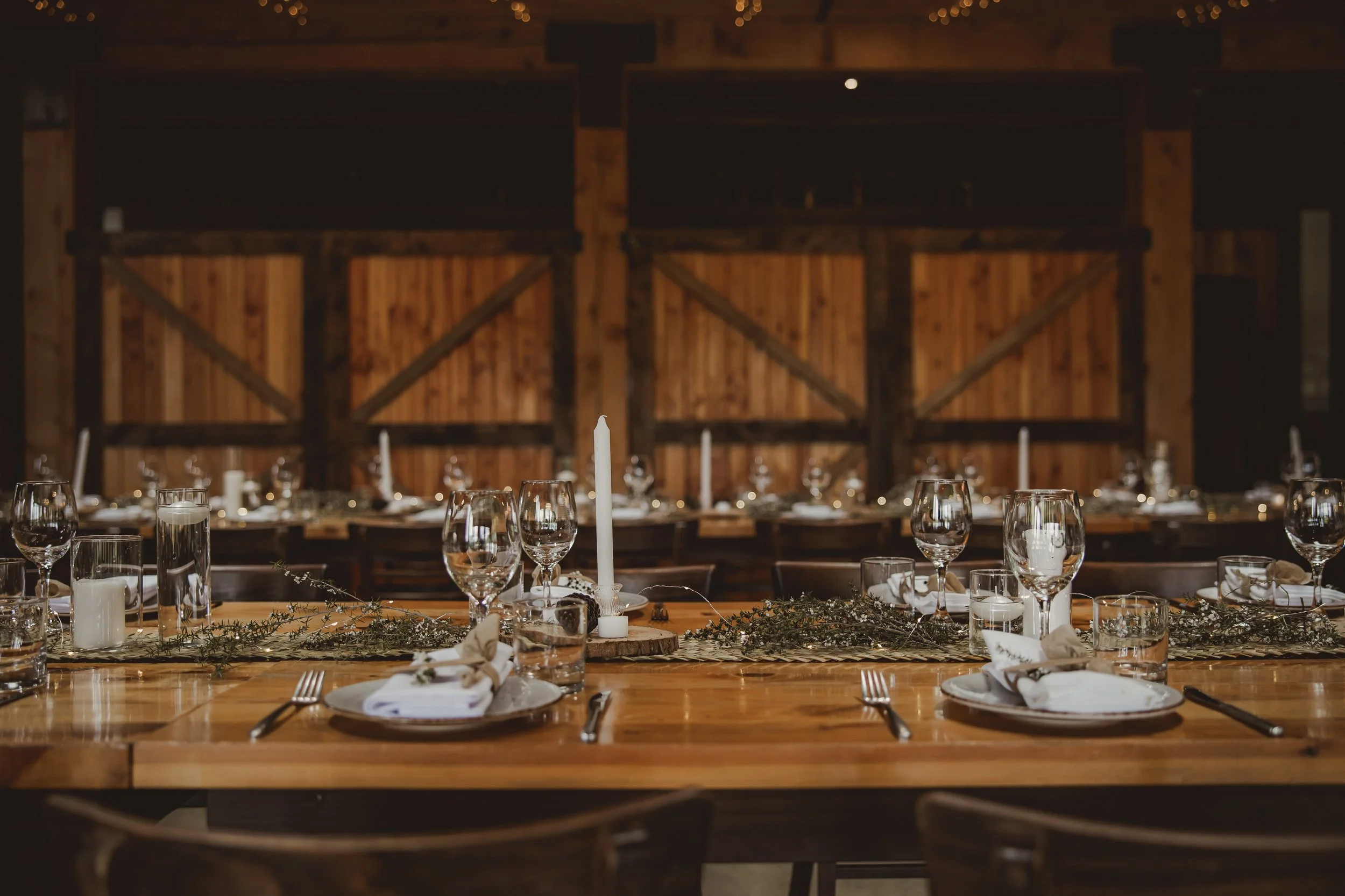 A rustic wooden banquet table set for a formal event with wine glasses, candles, plates, napkins, and decorative greenery, in a barn-like setting with wooden walls.