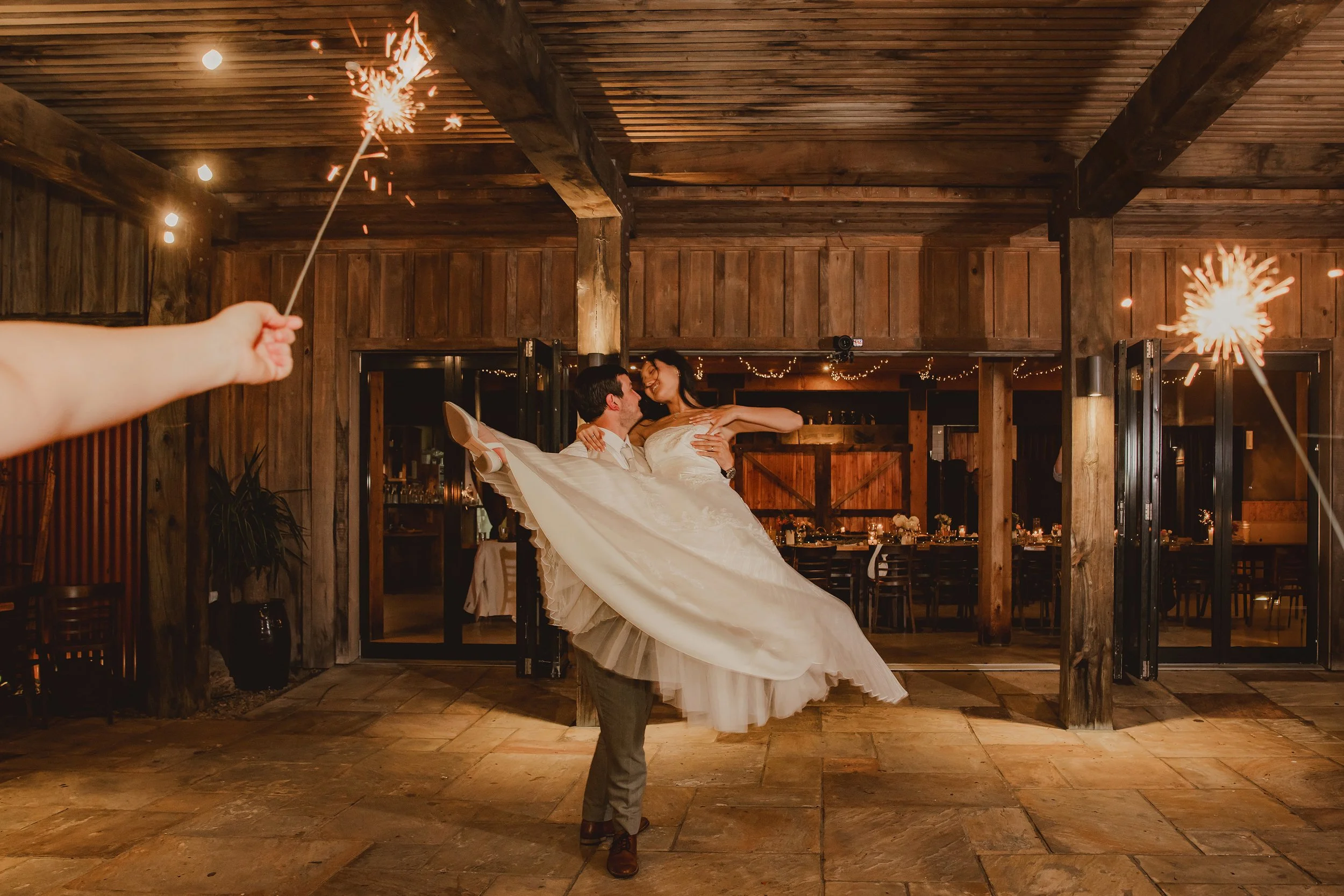 A couple dancing at their wedding reception, with the groom lifting the bride in a wooden venue, holding sparklers.