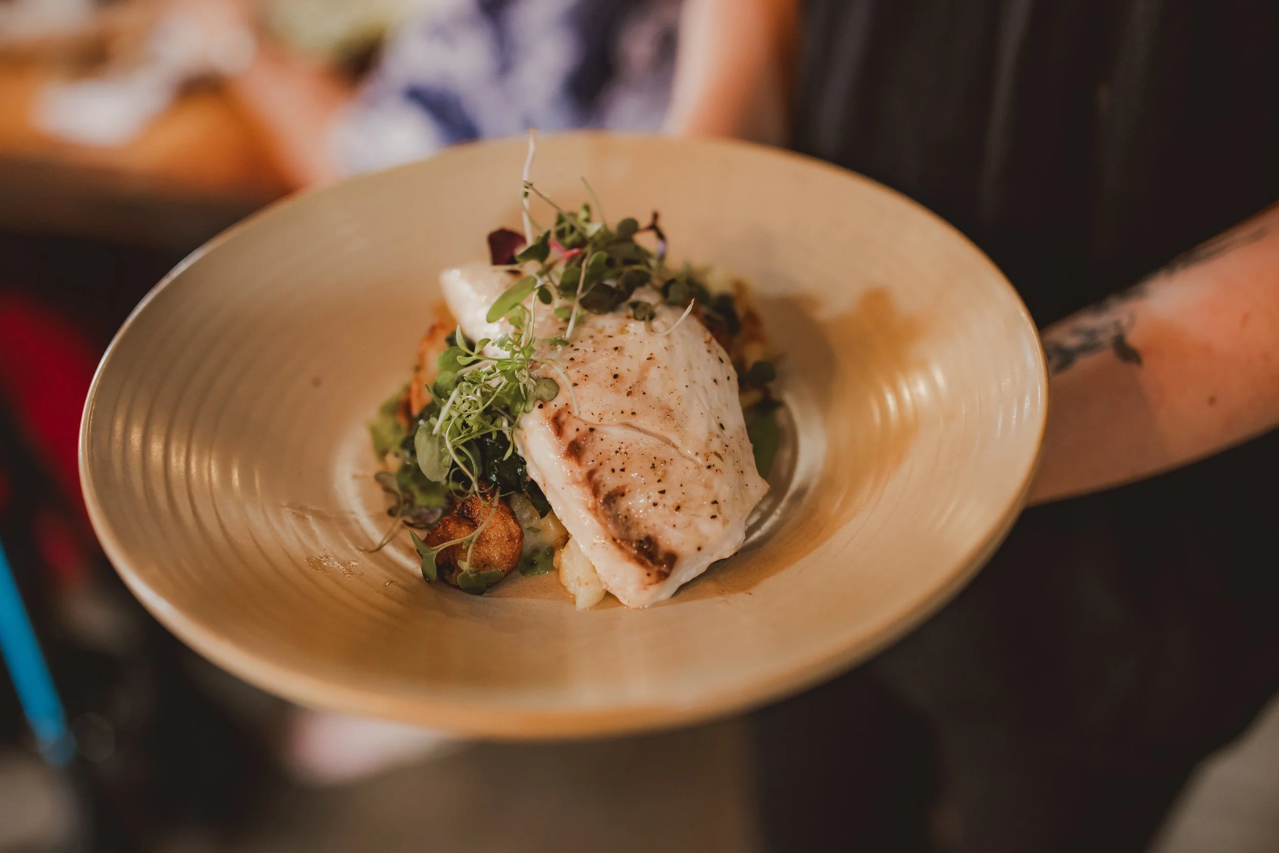 A plated dish featuring a piece of white fish, garnished with microgreens, served on a beige, ridged plate.