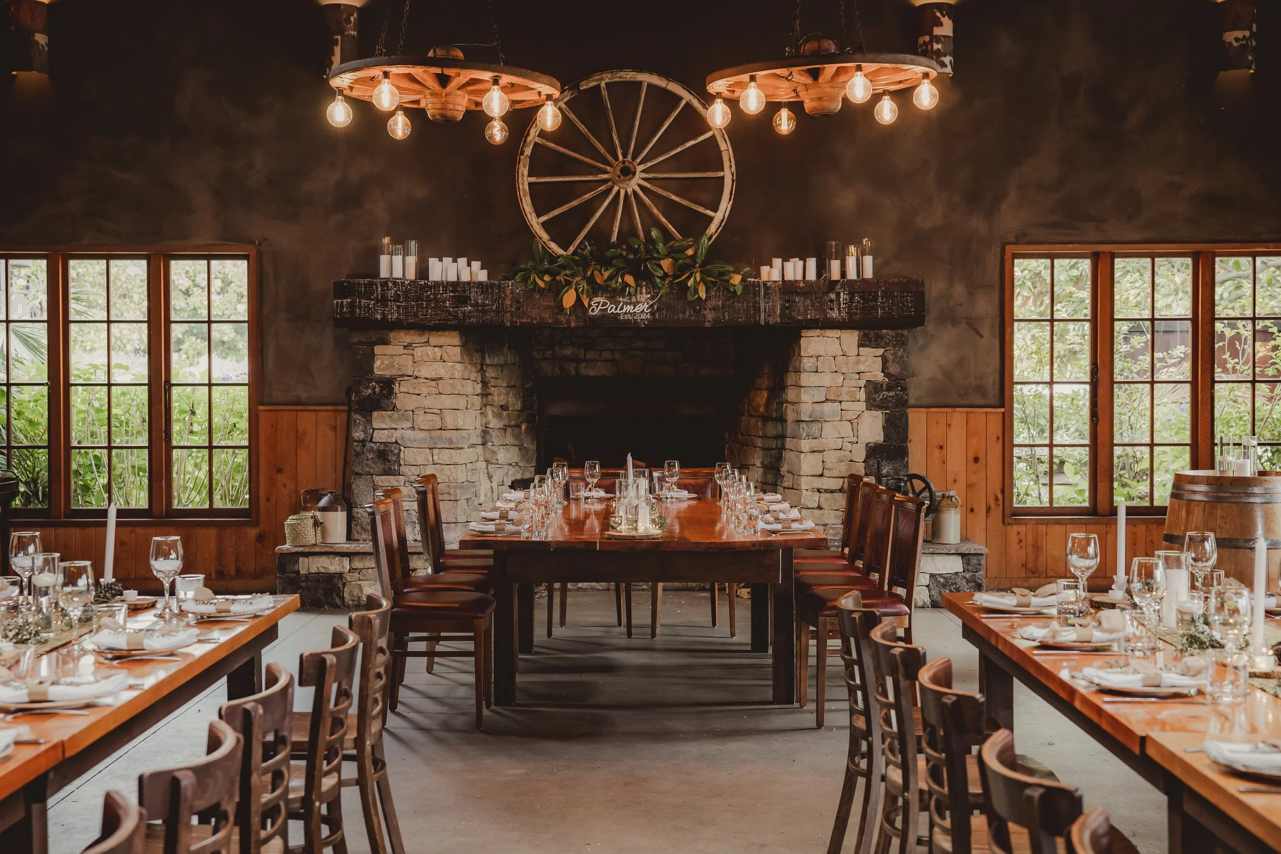 A rustic indoor dining area with long wooden tables set with glassware, plates, and napkins, arranged for a formal meal, in front of a stone fireplace with a wooden mantel decorated with candles and greenery, large windows on each side providing natu