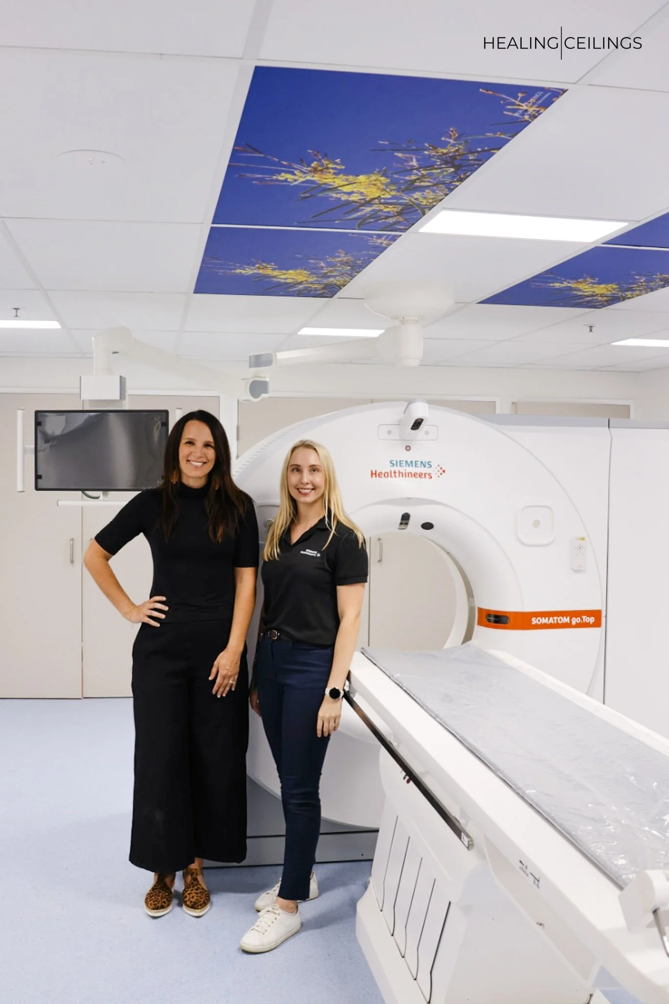 Two women standing next to an MRI machine in a medical imaging room, with a ceiling decorated with blue sky and yellow flowers designs.