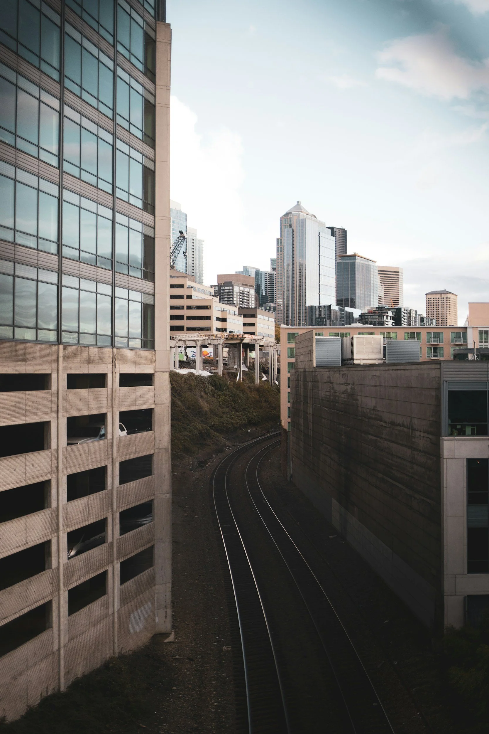 Urban cityscape with skyscrapers, railroad tracks curving between buildings, and a partly cloudy sky.