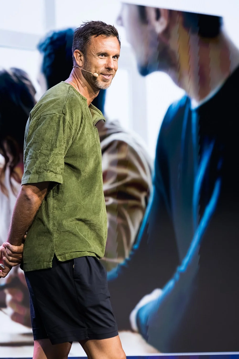 A casually dressed conference presenter smiles as he looks toward the audience