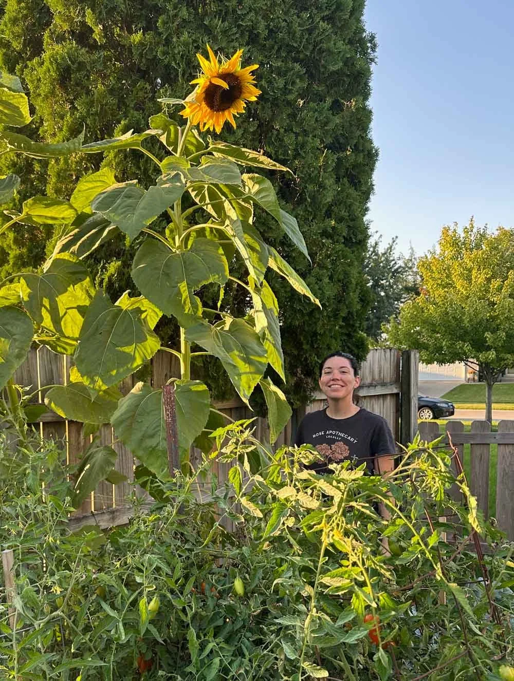 A woman smiling surrounded by her garden which includes tall sunflowers.