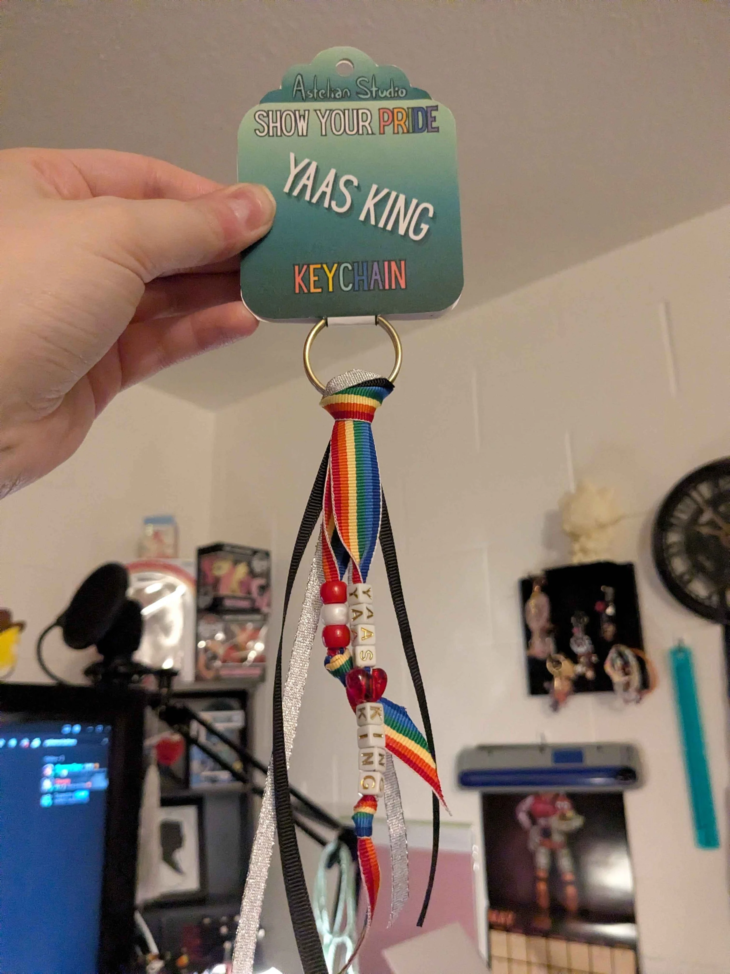 whole keychain detail that says Yaas King in beads. With rainbow, black and silver ribbon tassels against a blurry background of a computer room with white walls. The keychain is being held up by a person with light skin holding the product card.