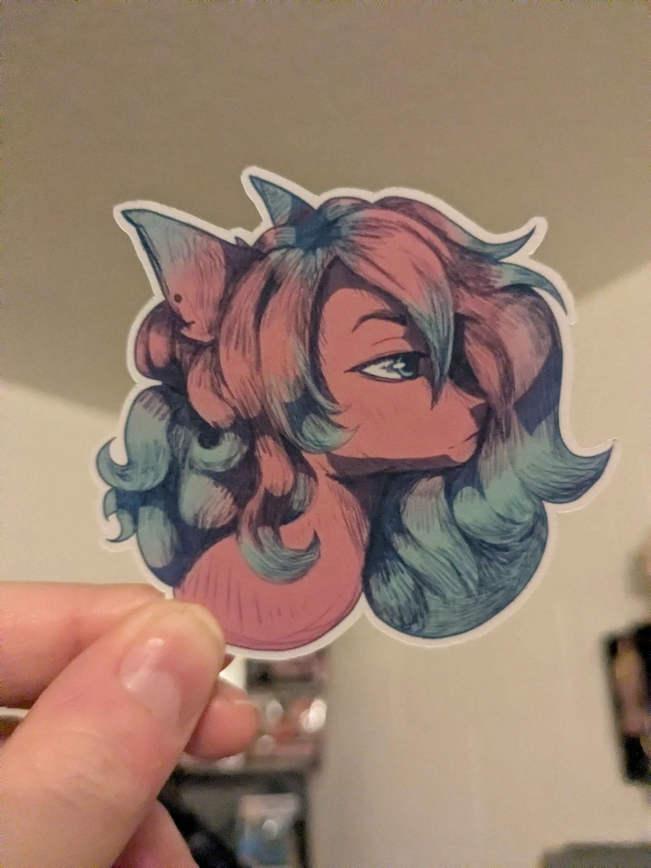 sticker behing held up against a blurry white wall by a light skinned hand. depicts a headshot of a cartoon horse made to resemble the my little pony, friendship is magic style. The colors are mostly muted pink with green around the ends of the mane