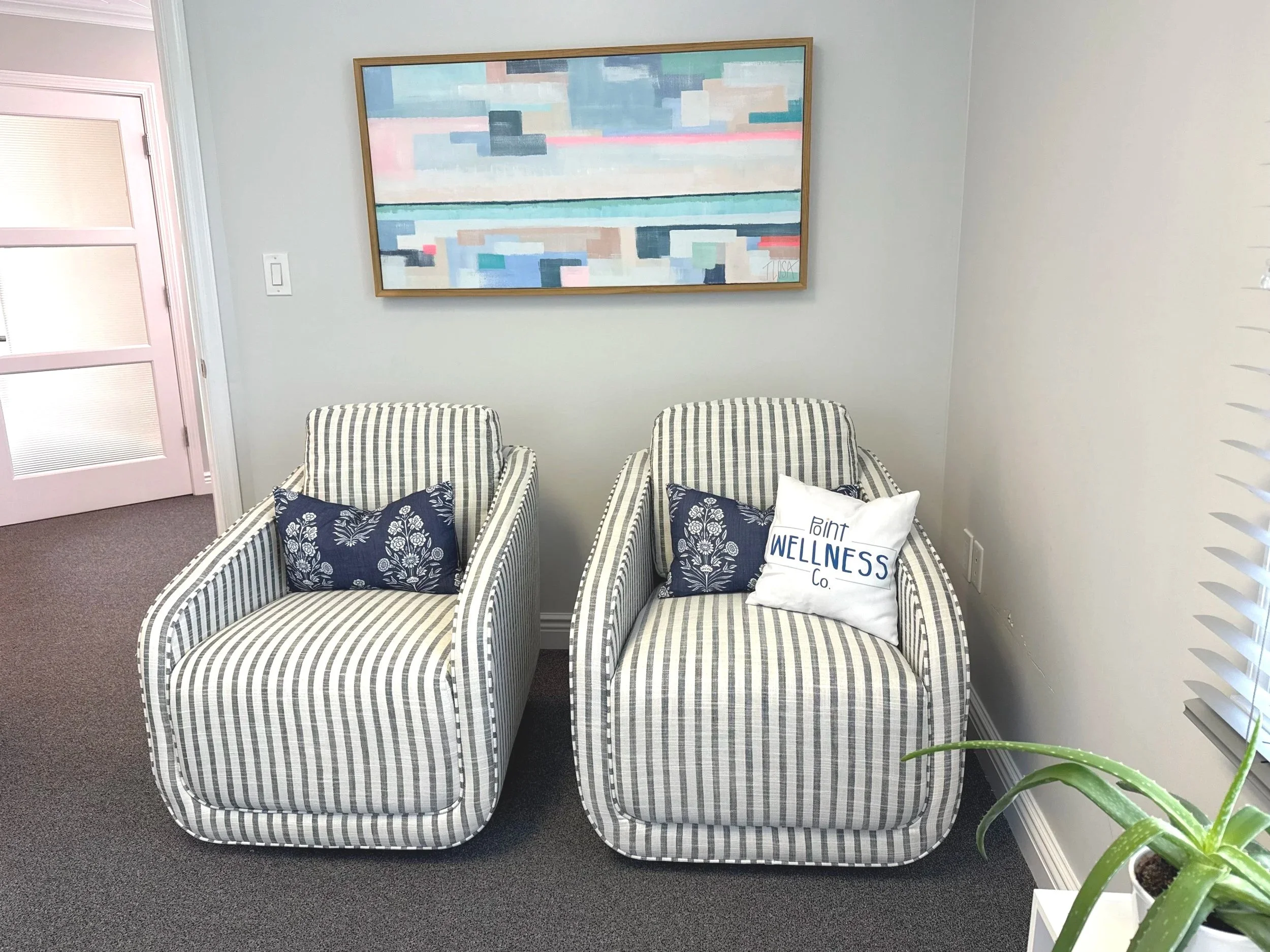 Two striped armchairs with decorative pillows in front of a light-colored wall with a large abstract painting. A potted plant is visible on the right in the foreground.