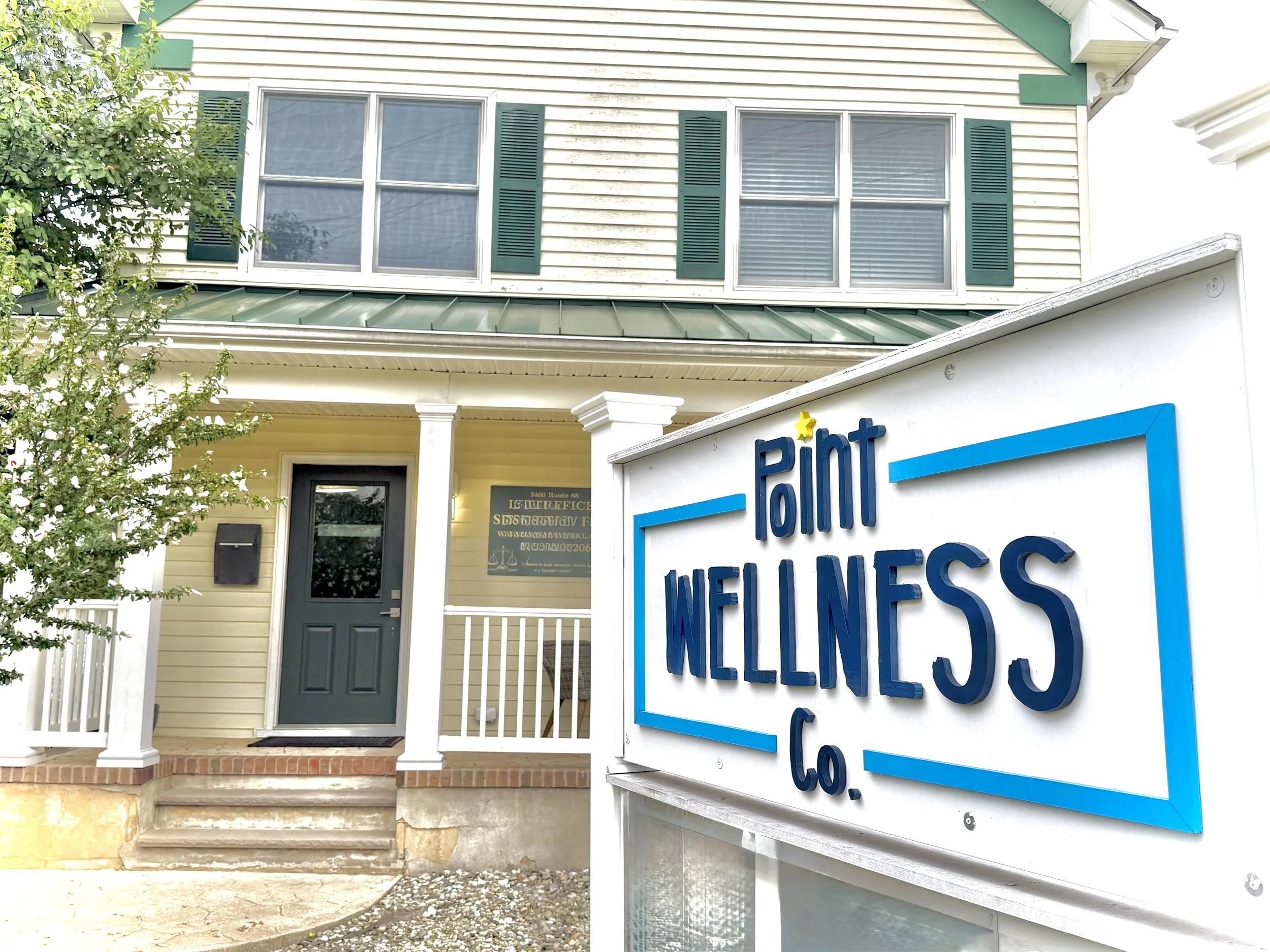 Exterior of a building with house siding and a sign reading 'Pint Wellness' in blue lettering with a yellow star, located near the entrance with steps and a black door, surrounded by a small white picket fence and bushy green tree.