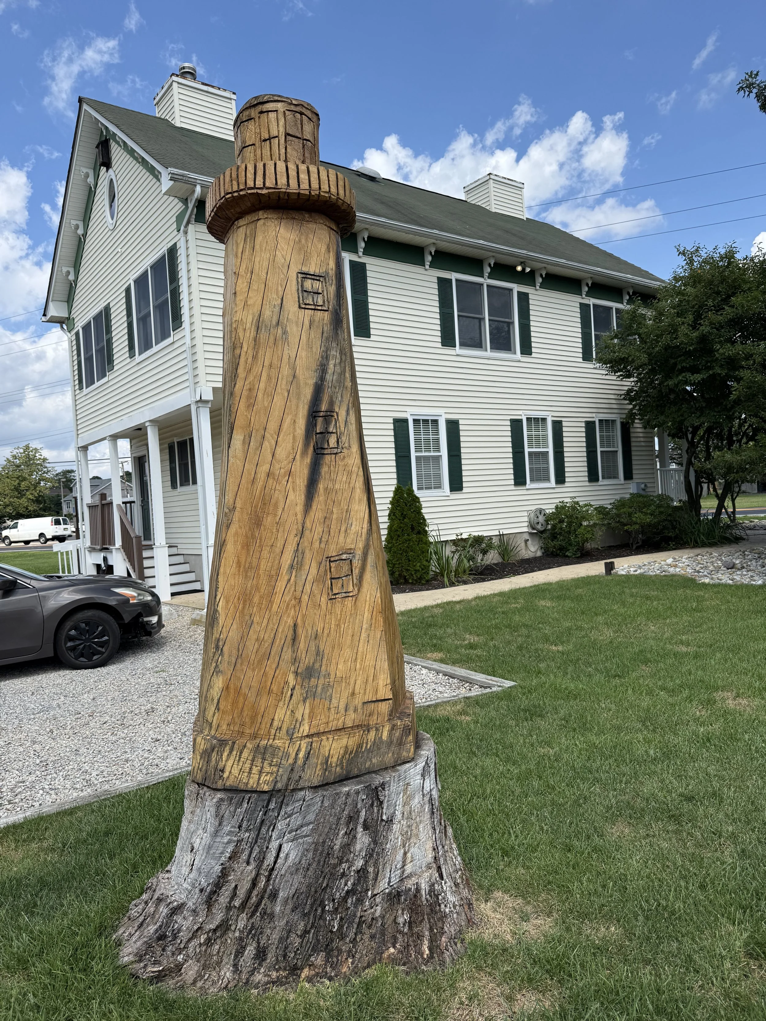 A large wooden chimney sculpture in front of a white house with green shutters, a gray shingle roof, and a chimney pipe, under a partly cloudy blue sky.