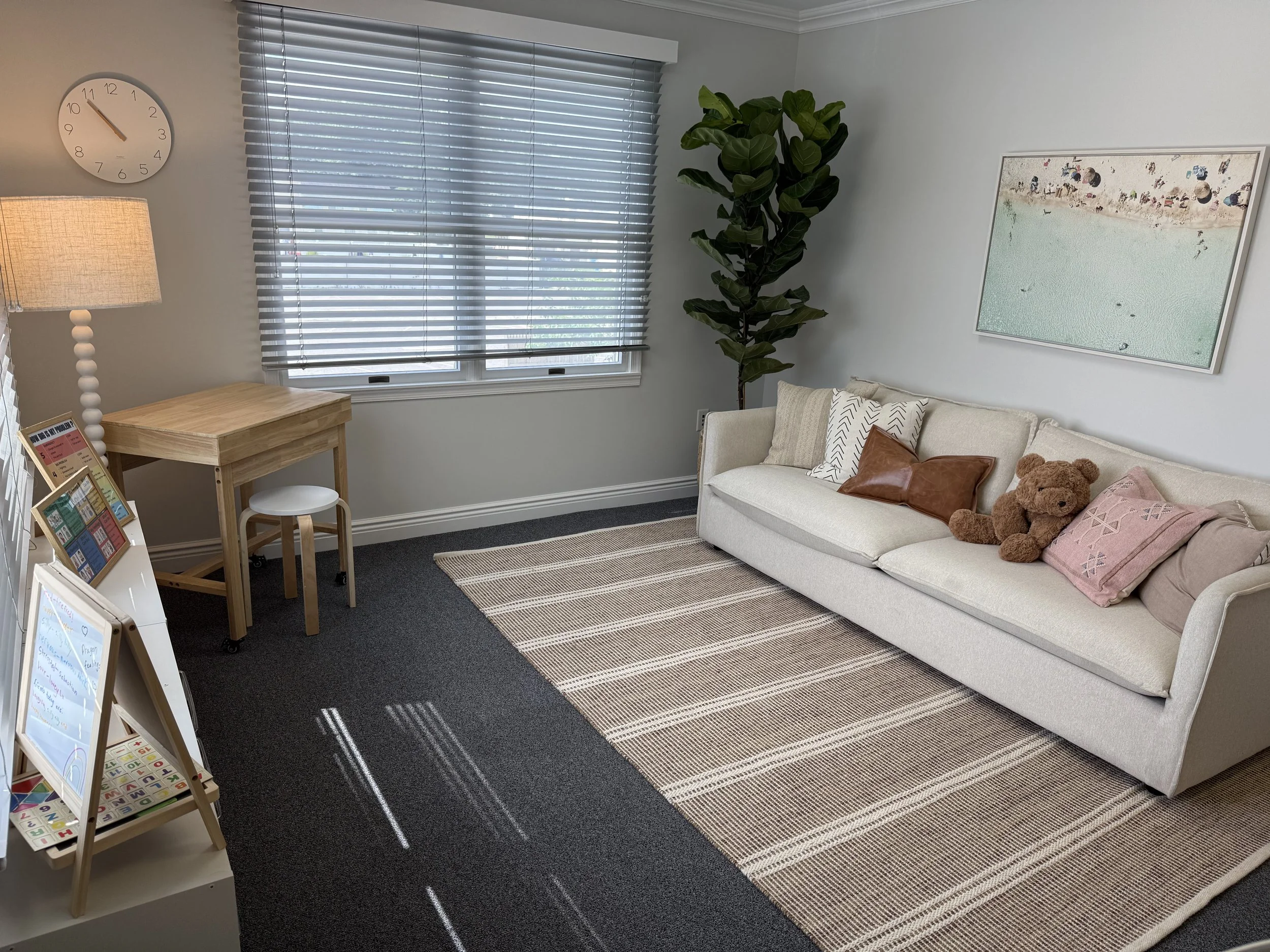 A living room corner with a beige sofa adorned with pillows, including one pink pillow and a teddy bear, a wooden side table with magazines, a floor lamp with a textured shade, a large green leafy plant in the corner, a window with horizontal blinds,