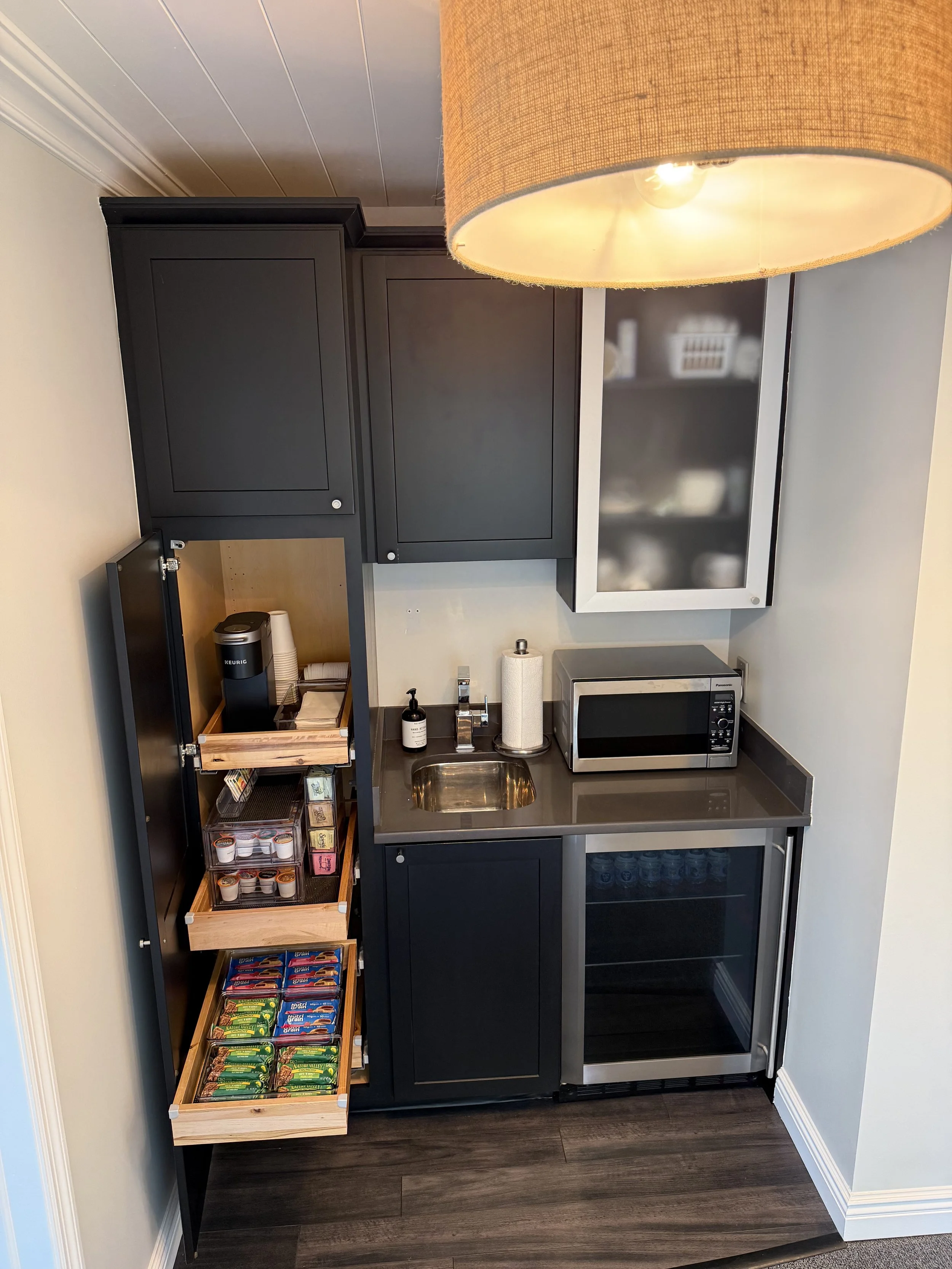 A kitchenette with a black cabinet, a microwave, a coffee maker, and snack racks with various snack packages. There is a large lamp overhead.