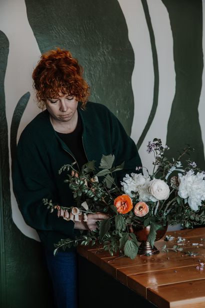 Woman arranging a floral arrangement of white, orange, and purple flowers on a wooden table.