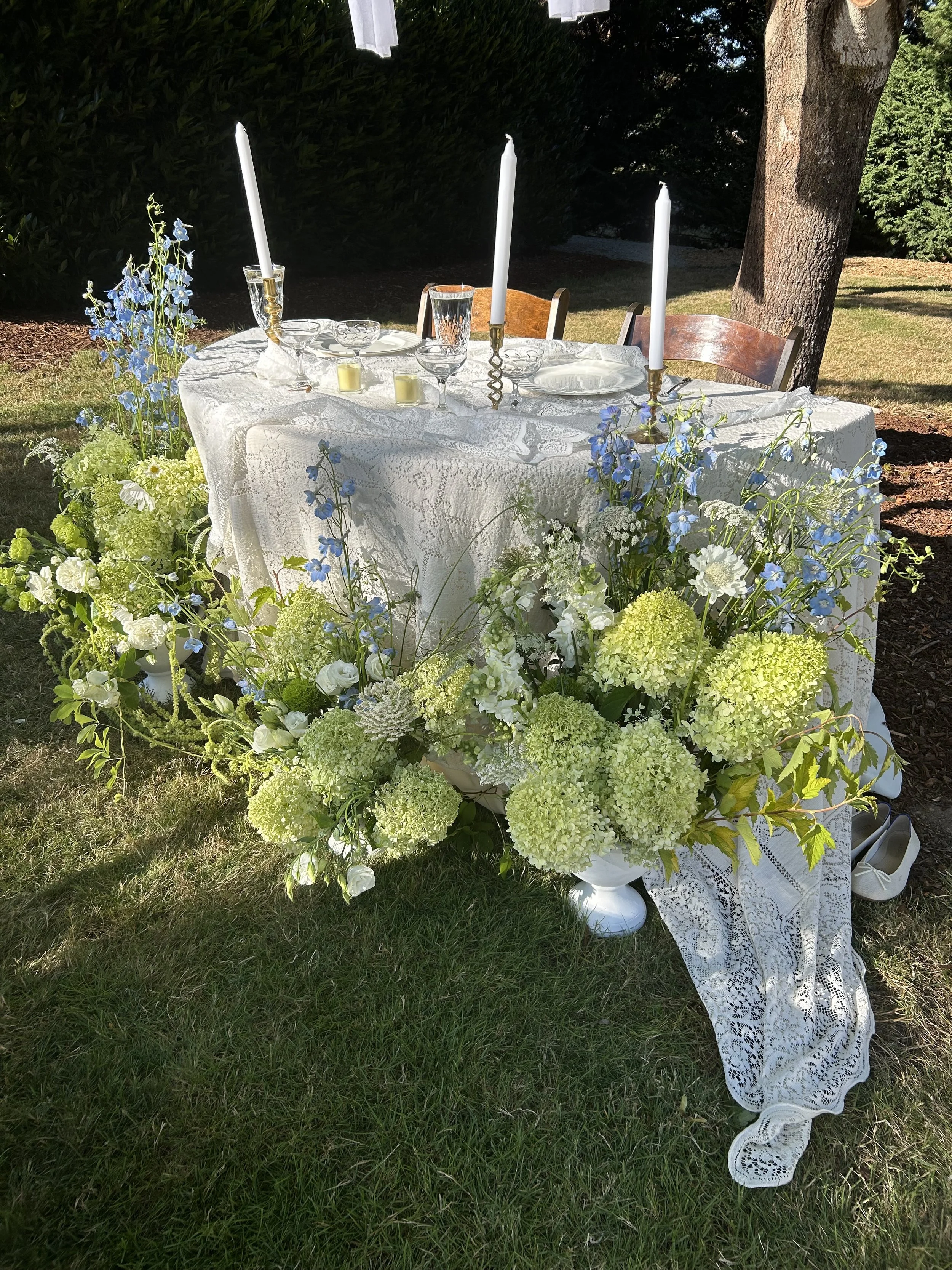 A long table covered with a white lace tablecloth is decorated with blue and white flowers, candles, and glassware, set outdoors beneath a tree.