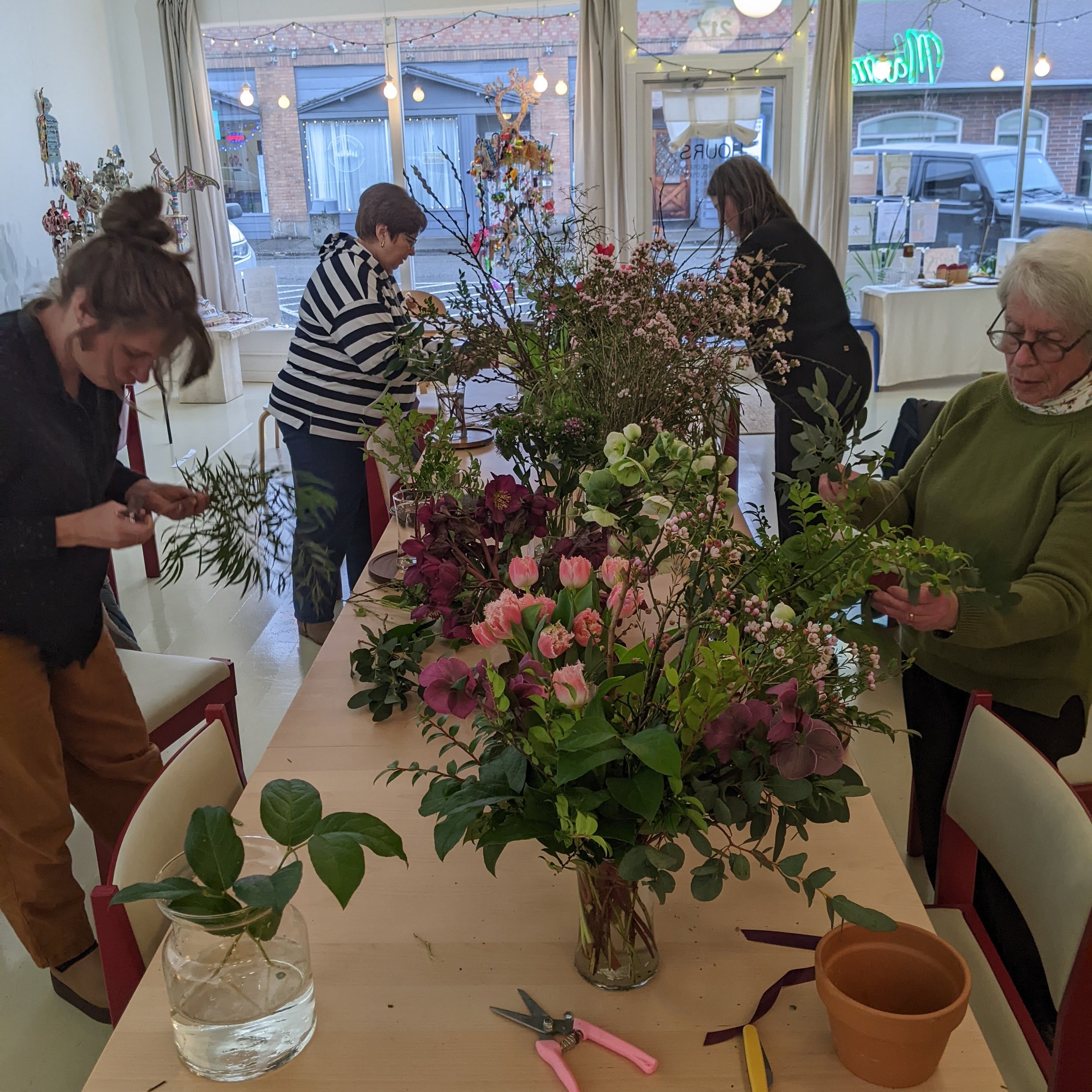 Interior view of a flower shop with customers arranging flowers on a table; large vases with pink and green flowers, scissors, and pots are visible; outside seen through large windows showing a street with buildings and vehicles; decorative lights hang from the ceiling.