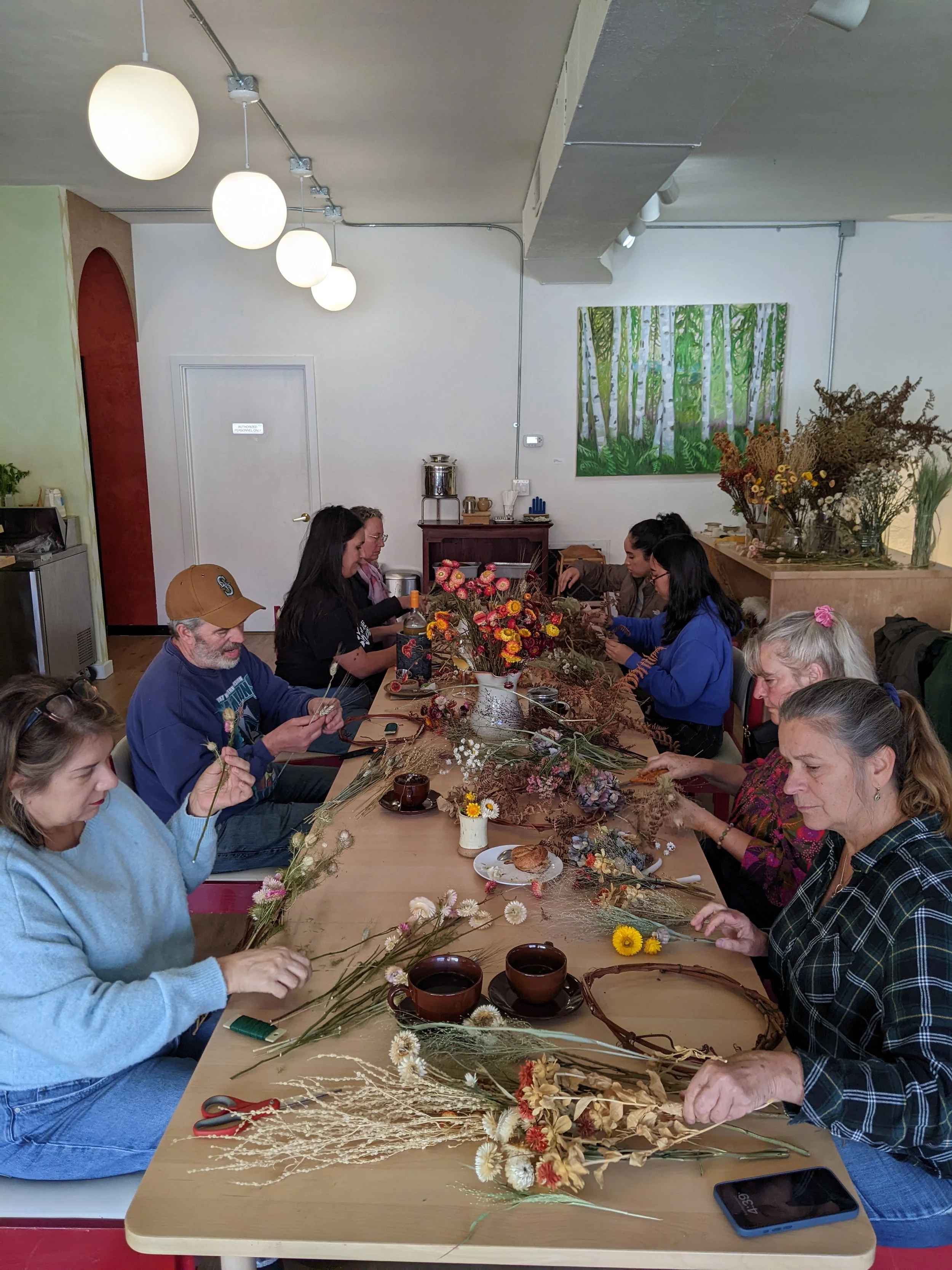 People sitting at a table creating floral arrangements with dried flowers, surrounded by various tools and supplies in a room decorated with artwork and flowers.
