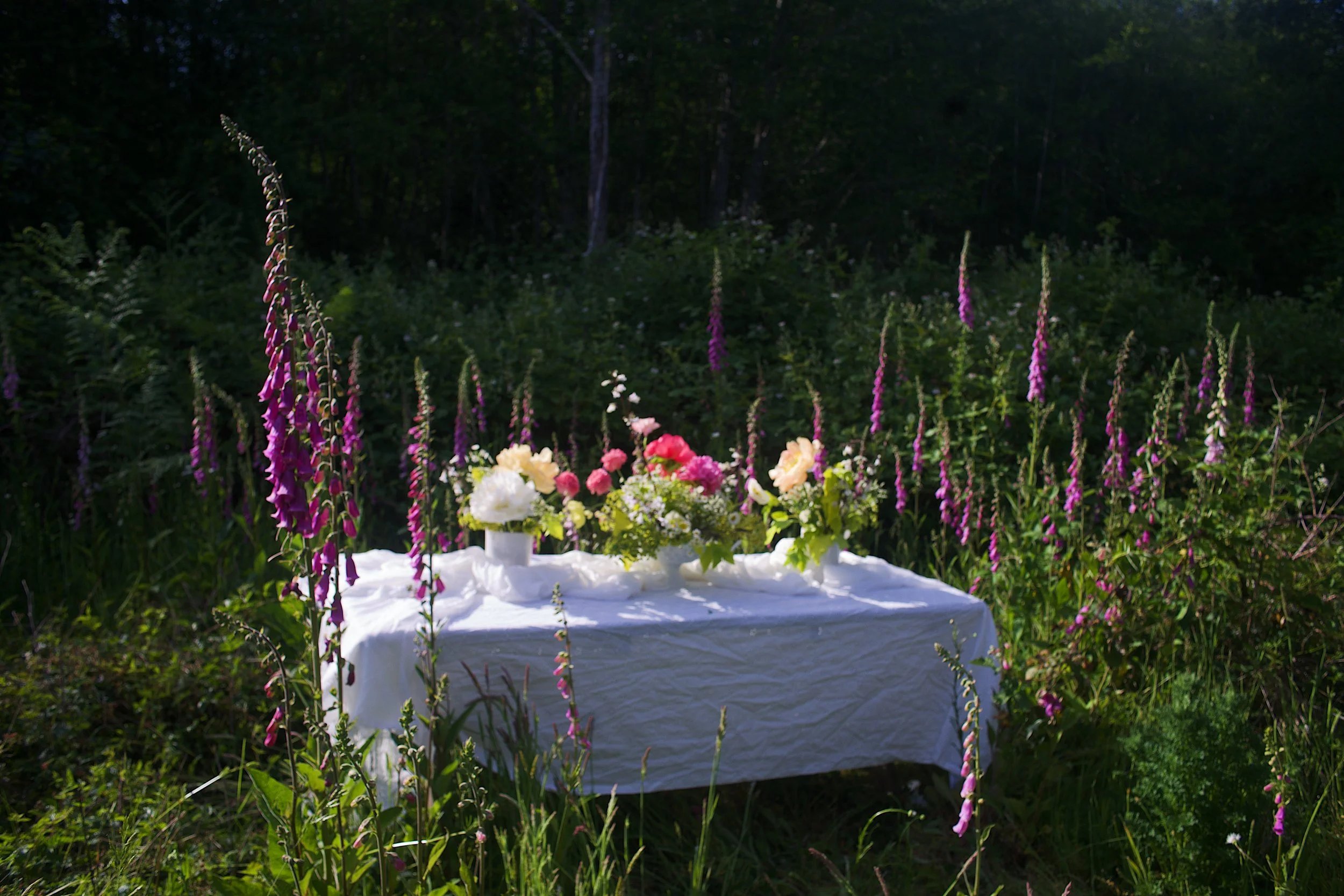 A table with a white cloth is set outdoors in a lush garden, decorated with a large floral arrangement of pink, white, and yellow flowers. Tall purple foxgloves surround the table, with a dense wooded area in the background.