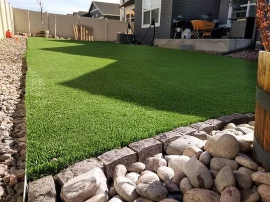 A backyard with a well-maintained artificial grass lawn, bordered by a line of small rocks and larger decorative stones. In the background, there is a house with a deck, a wheelbarrow, and some gardening tools.