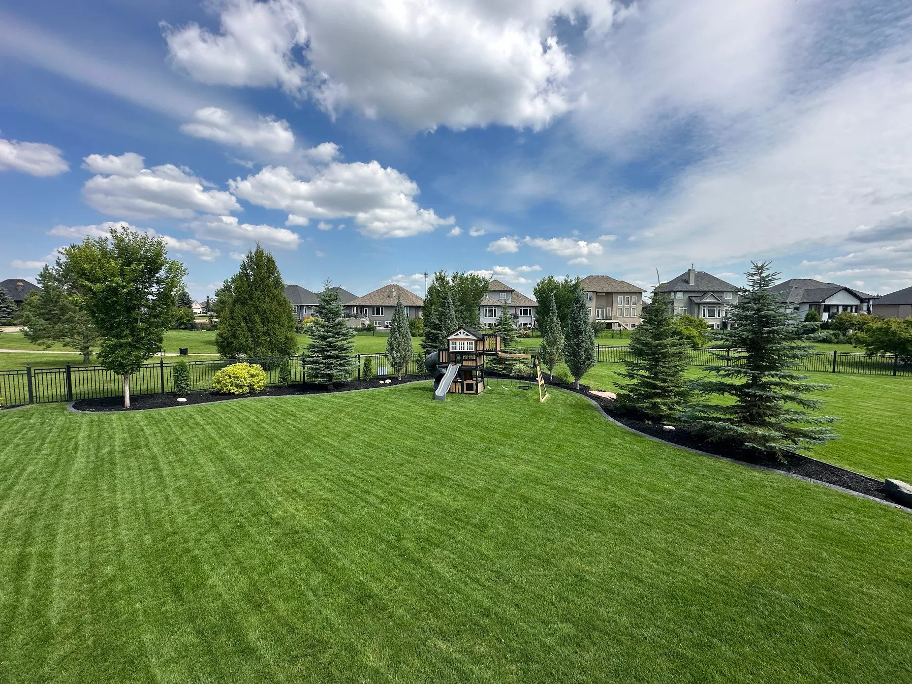 A backyard with a well-maintained green lawn, a children's playset with slide and swings, surrounded by a black fence. There are flowers and trees along the fence, and houses with gray and white exteriors are visible in the background. The sky is partly cloudy with fluffy white clouds and patches of blue.