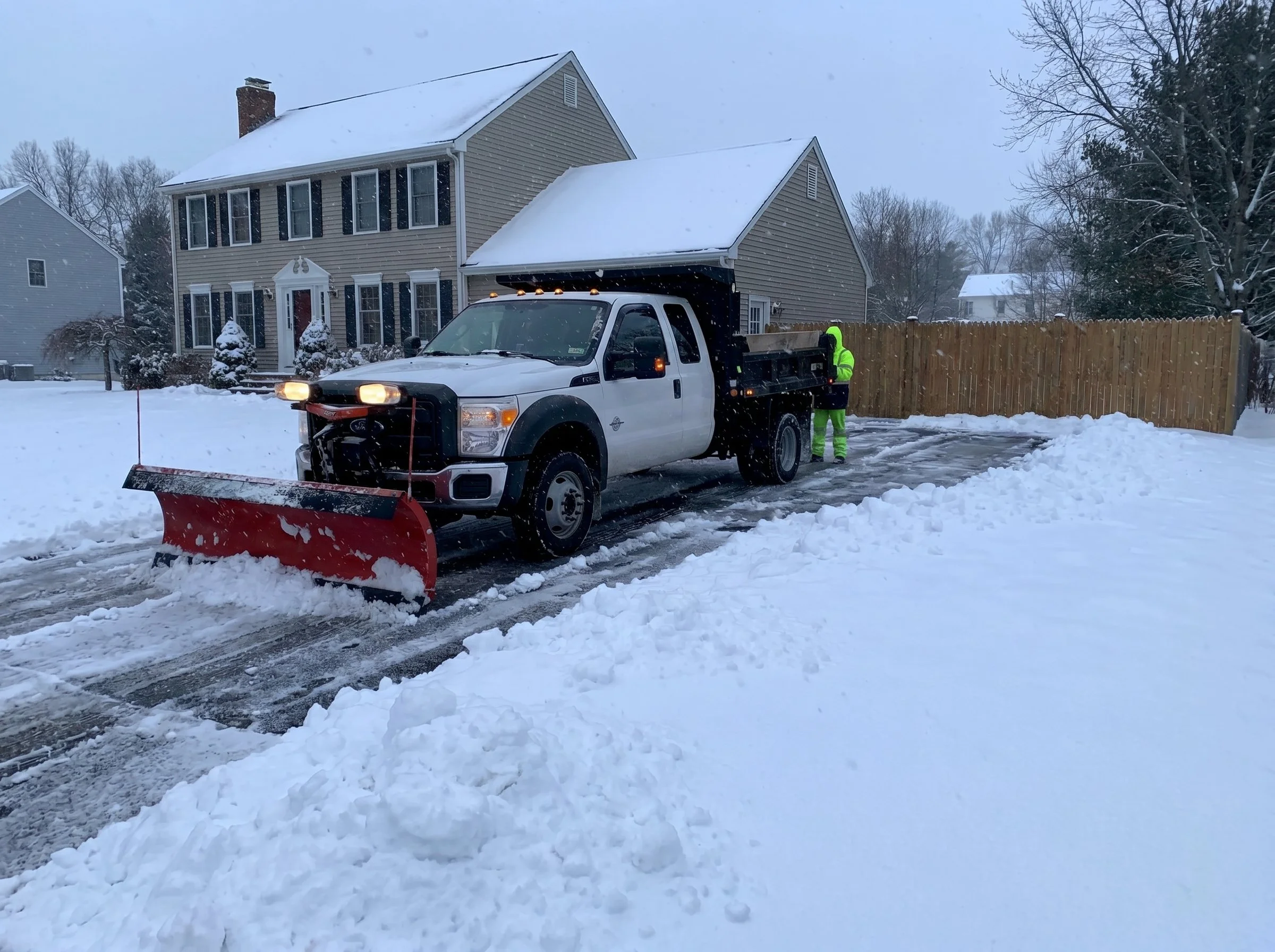A snow-covered residential street with a large house in the background and a snowplow truck clearing the road, with a worker in reflective clothing nearby.