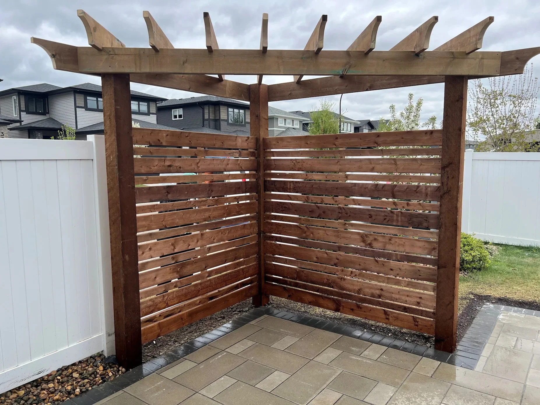 A partially constructed outdoor wooden privacy screen with slatted panels and a horizontal top beam, situated on a patio with a tiled floor, in a residential backyard with neighboring houses visible in the background under cloudy skies.
