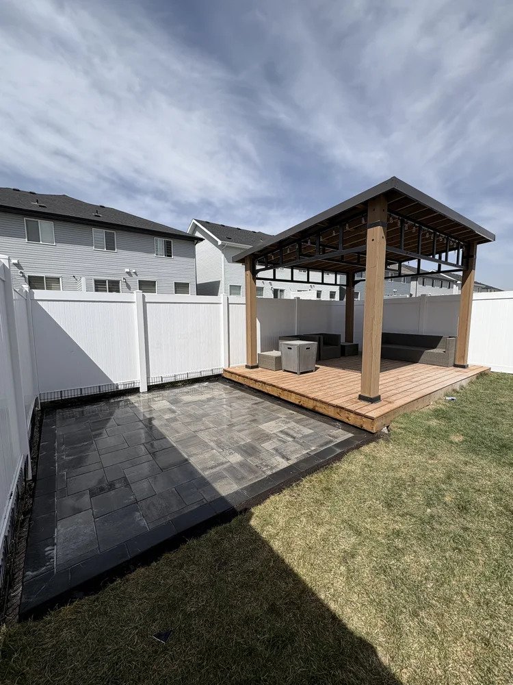 A backyard with a white privacy fence, a patio with dark gray paving stones, and a wooden deck with a pergola. The deck has seating and small tables. The sky is partly cloudy.