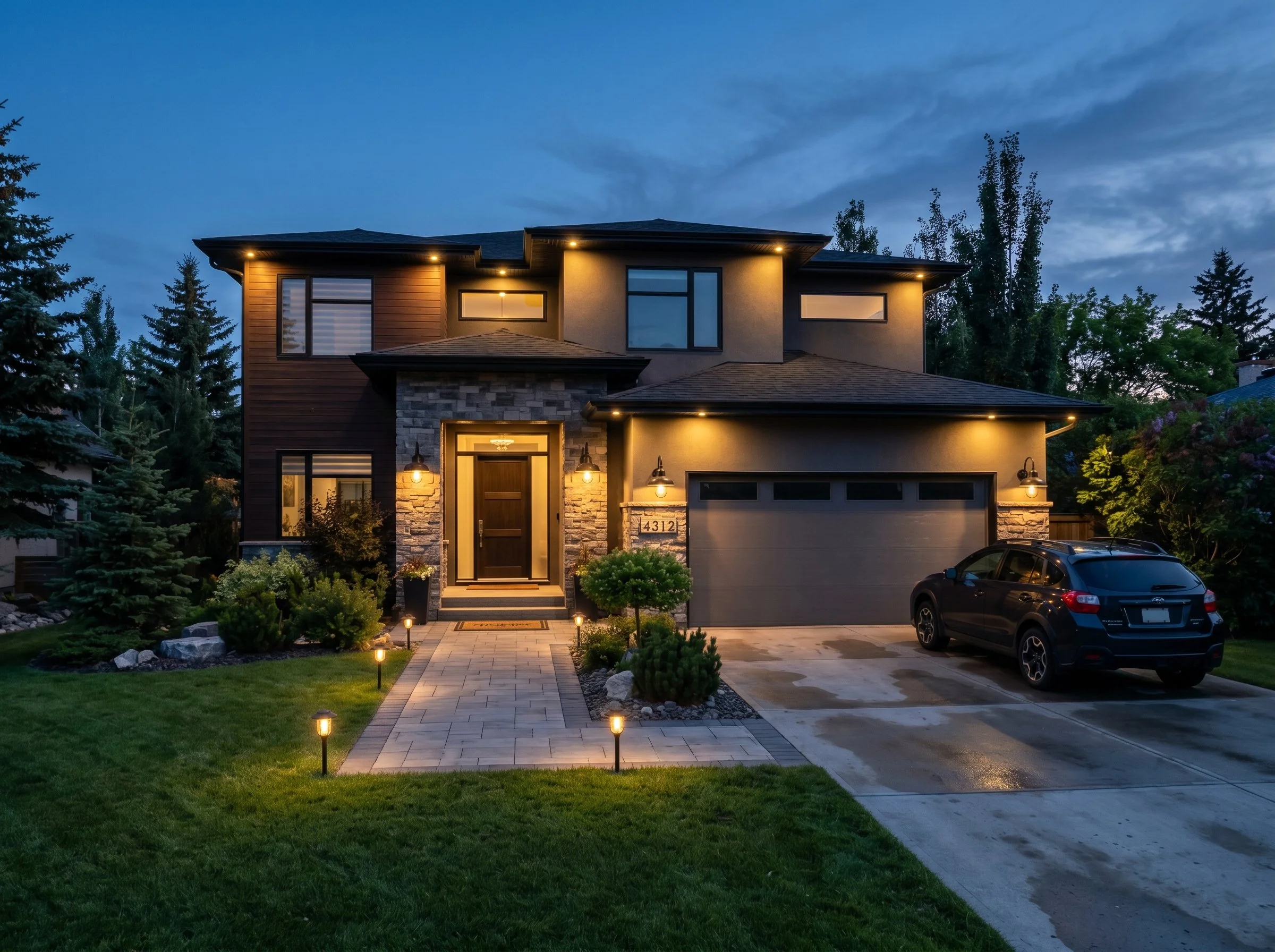 A two-story modern house illuminated at dusk, featuring a paver driveway, a landscaped front yard with grass, small trees, and outdoor lighting. The house has a mix of stone and wood exterior, large windows, and a garage with a black car parked outside.