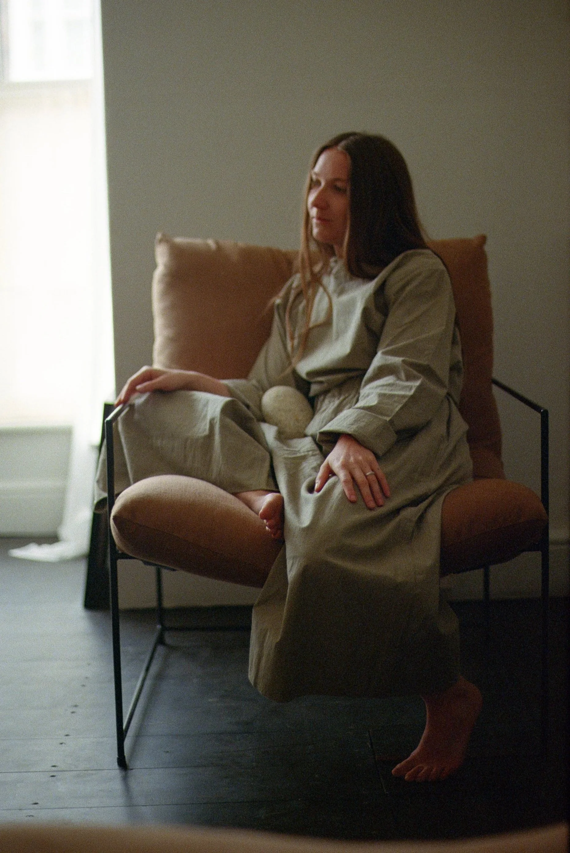 A woman sitting on a brown armchair with a neutral expression, dressed in a beige robe, in a softly lit room with a gray wall and a window with sheer curtains.