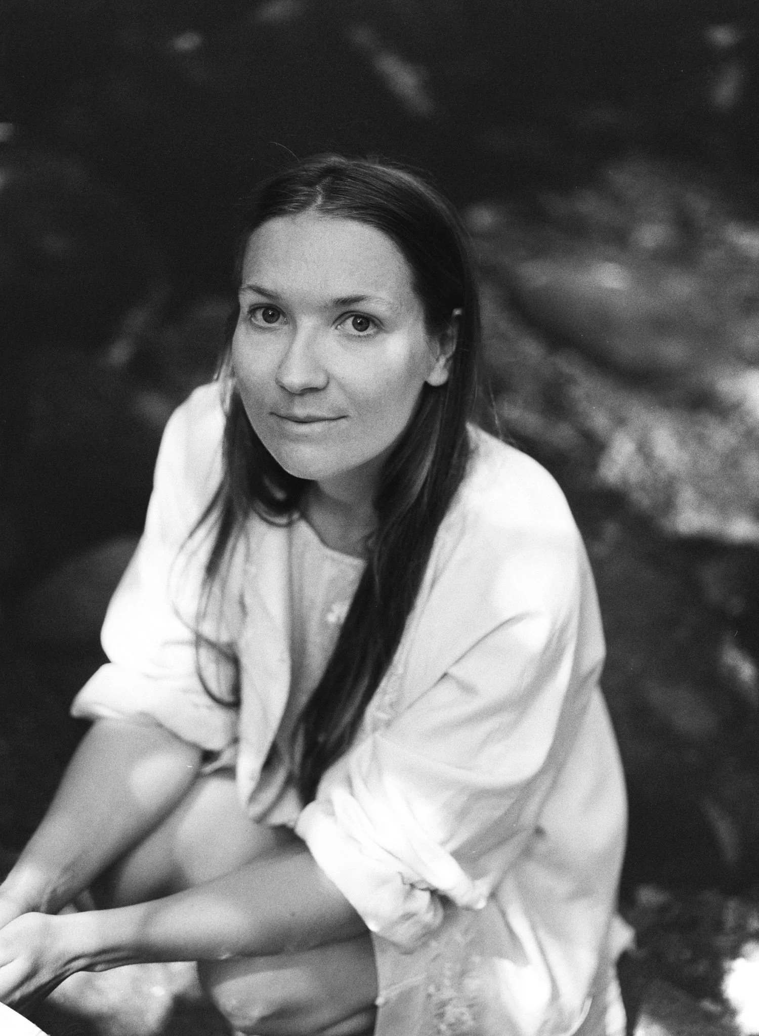 A black and white photo of a woman with long dark hair, wearing a white shirt, sitting outdoors near water, looking directly at the camera with a slight smile.