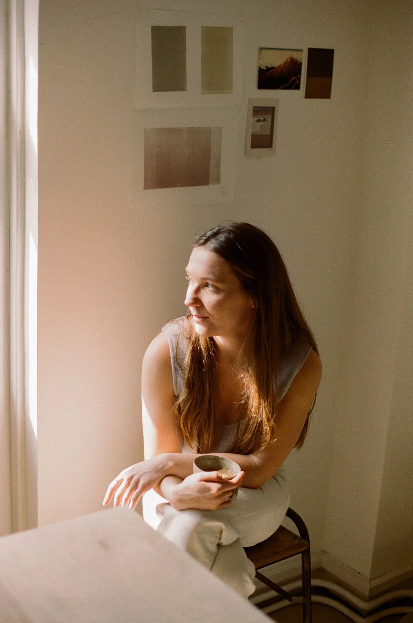 A woman with long brown hair sitting on a wooden chair in a room, holding a small bowl, looking thoughtfully out of a window, with sunlight illuminating her face. Wall art hanging behind her.
