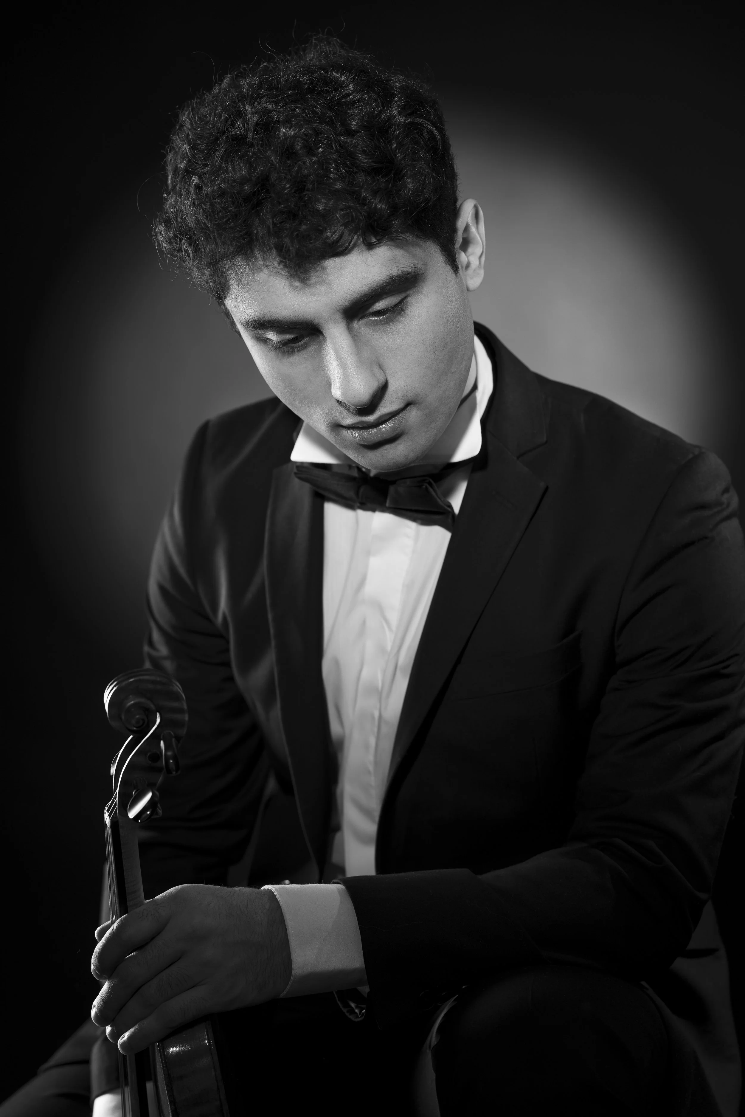 Svart-hvitt bilde av en ung mann i formell dress og sløyfe, som sitter med en fiolin i hånden, ser ned og har et konsentrert uttrykk.