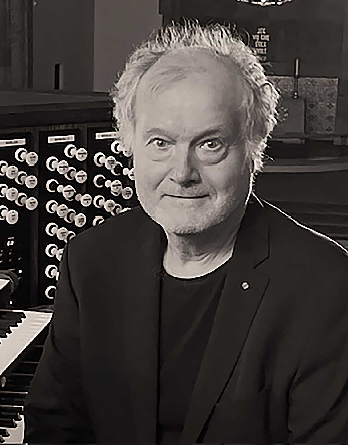 Et svart-hvitt bilde av en eldre mann med hvitt, krøllete hår, iført svart blazer, sittende ved en piano med musikkormer i bakgrunnen.