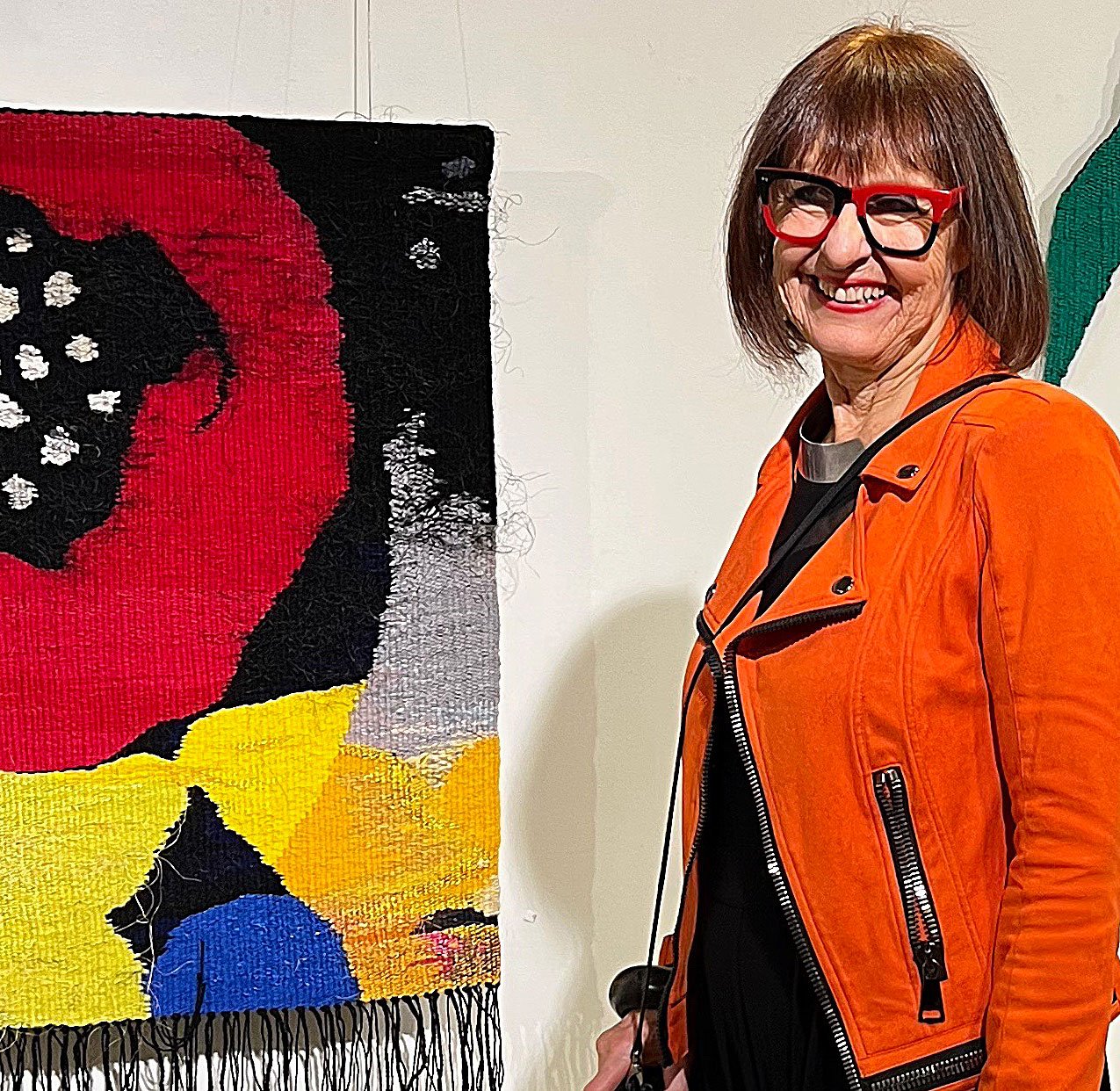 Kvinne med brunt hår, rød og svart rammet briller, iført oransje skinnjakke, smilende foran et kolorert kunstverk.