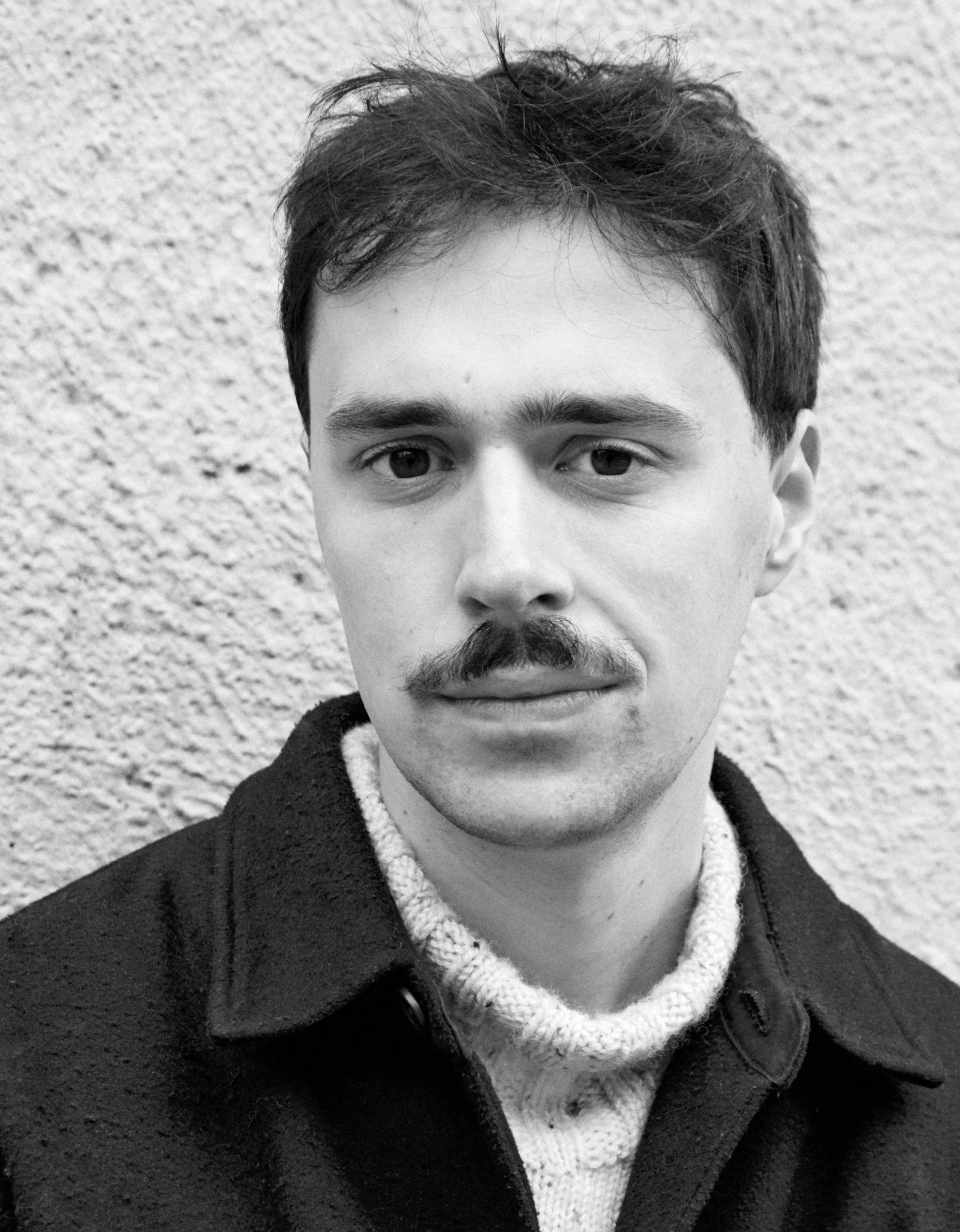 Sjenert ung mann med mørkt, kort hår og tykk bart, iført en mørk jakke og lys genser, foran en teksturert vegg.