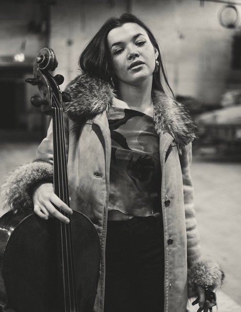 En kvinne holder en cello, iført en jakke med pelsdetaljer, i svart-hvitt bilde.