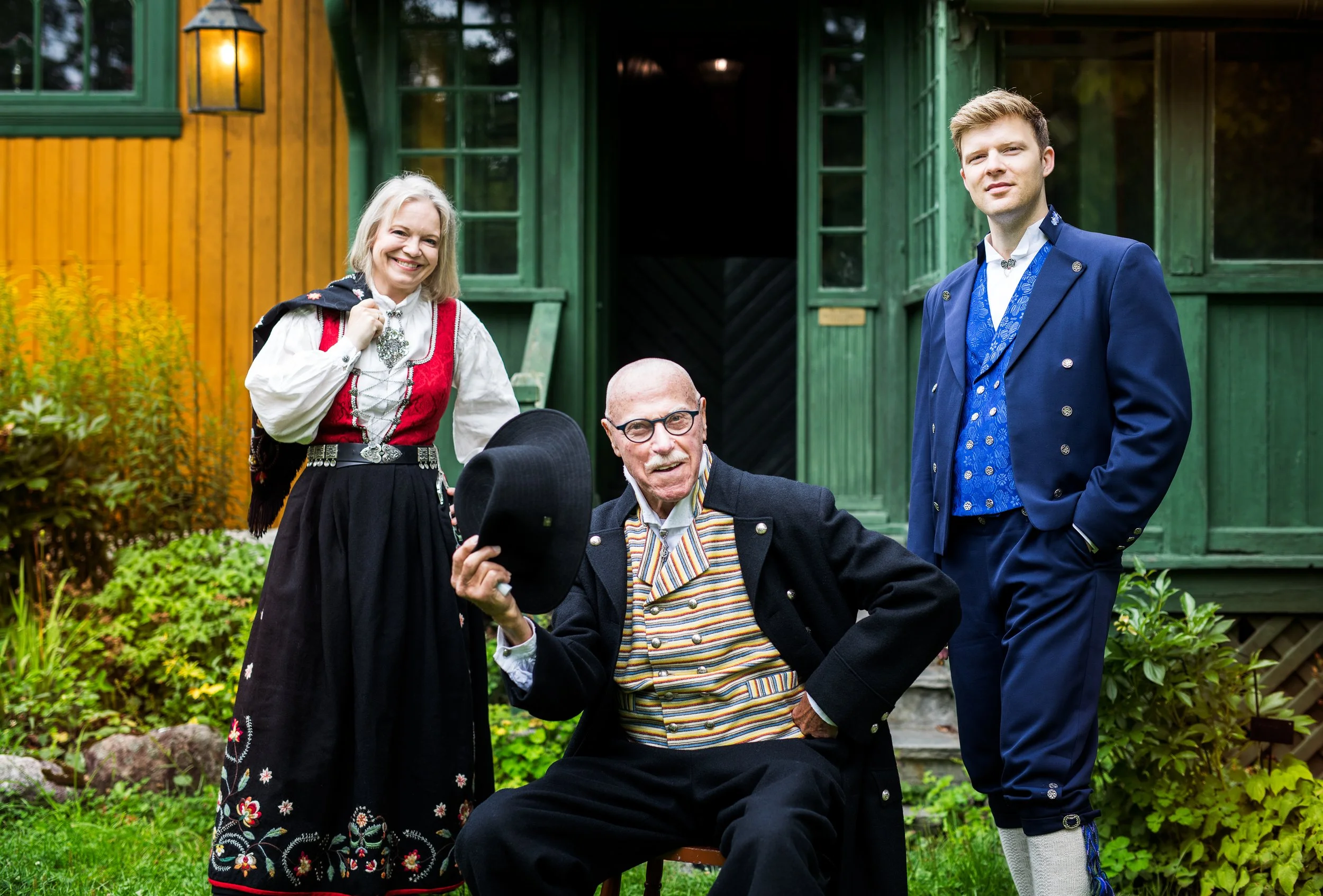 Fire mennesker iført tradisjonelle norske klær foran en fargesterk hytte, den eldste mannen med briller som holder en hatt, de andre smiler.